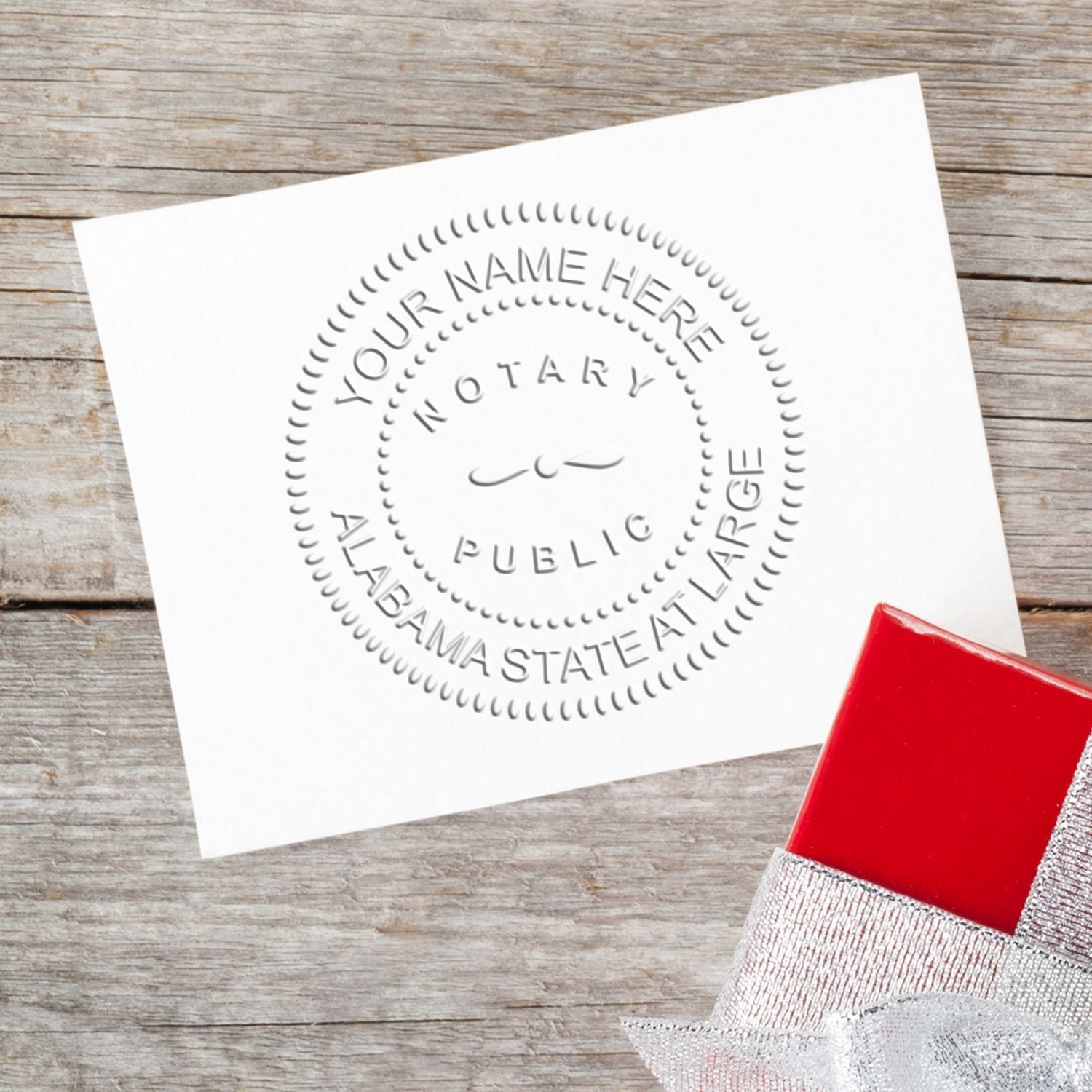 Embossed notary seal on white paper with a red gift box on a wooden surface. Blog post: The Roadmap to Notary Success: Alabama Notary Public Handbook Feature Image.