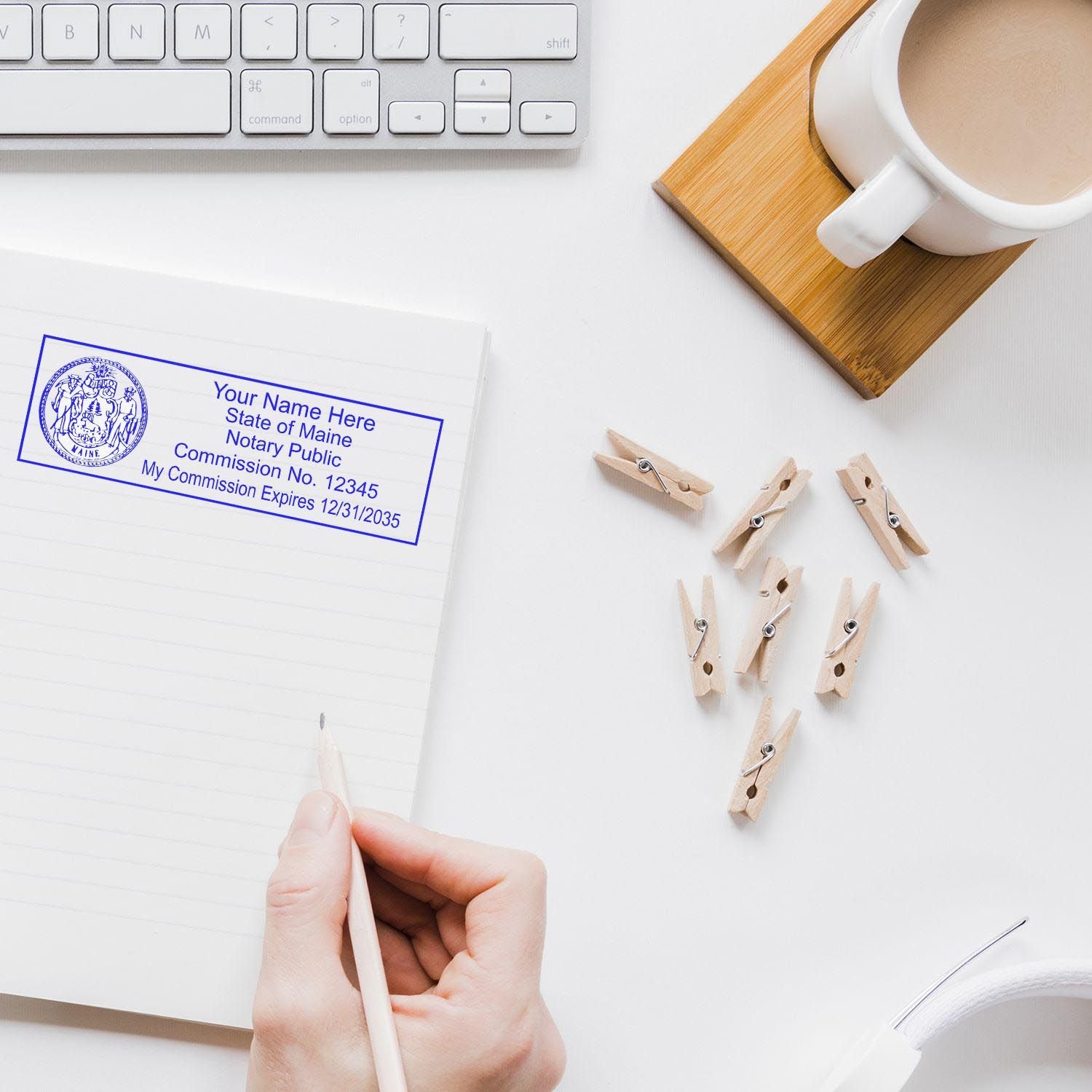 Maine Notary Supplies Blog Post Feature Image: A hand writing in a notebook with a Maine notary stamp, keyboard, coffee, and clothespins nearby.