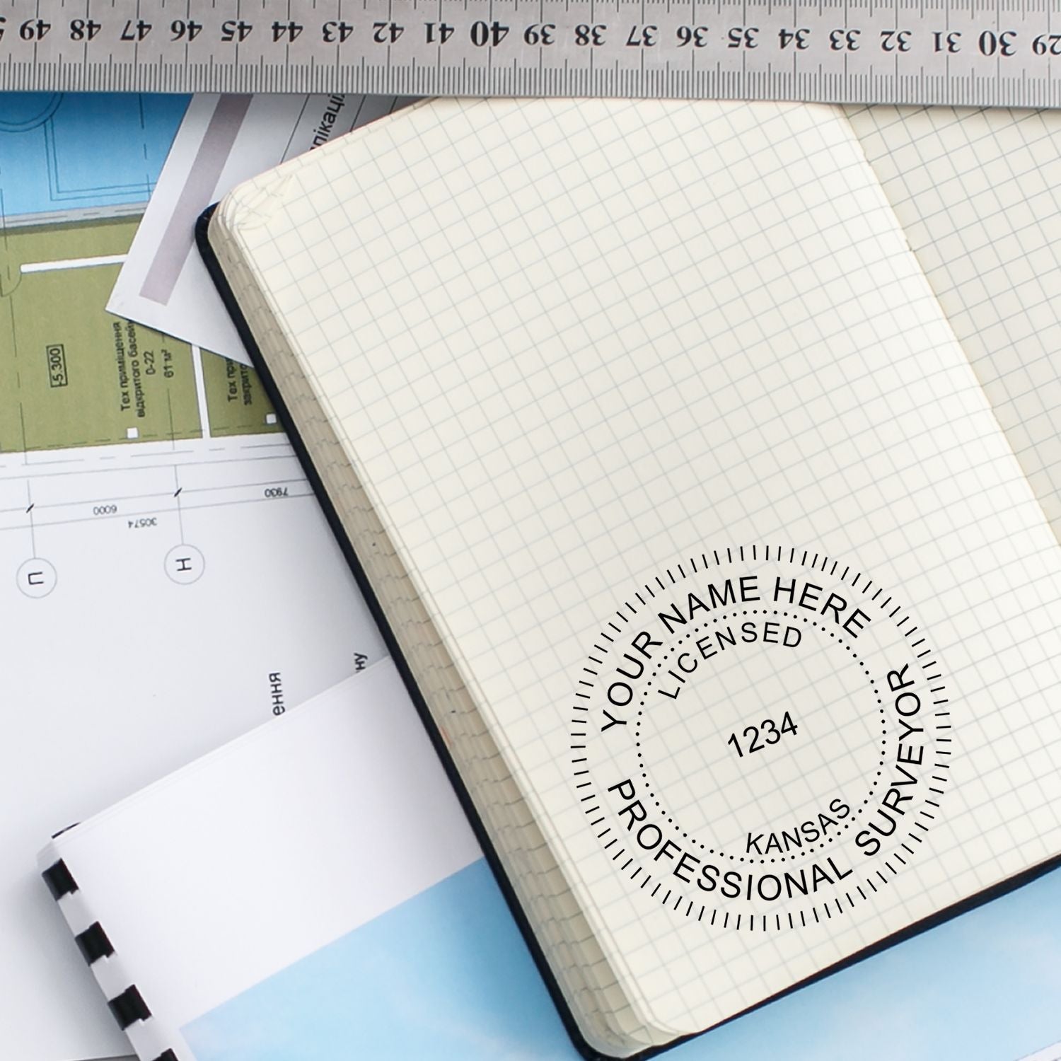 A Kansas Land Surveyor Seal stamp on a grid notebook, with architectural plans and a ruler in the background. Blog post: Kansas Land Surveyor Seal.