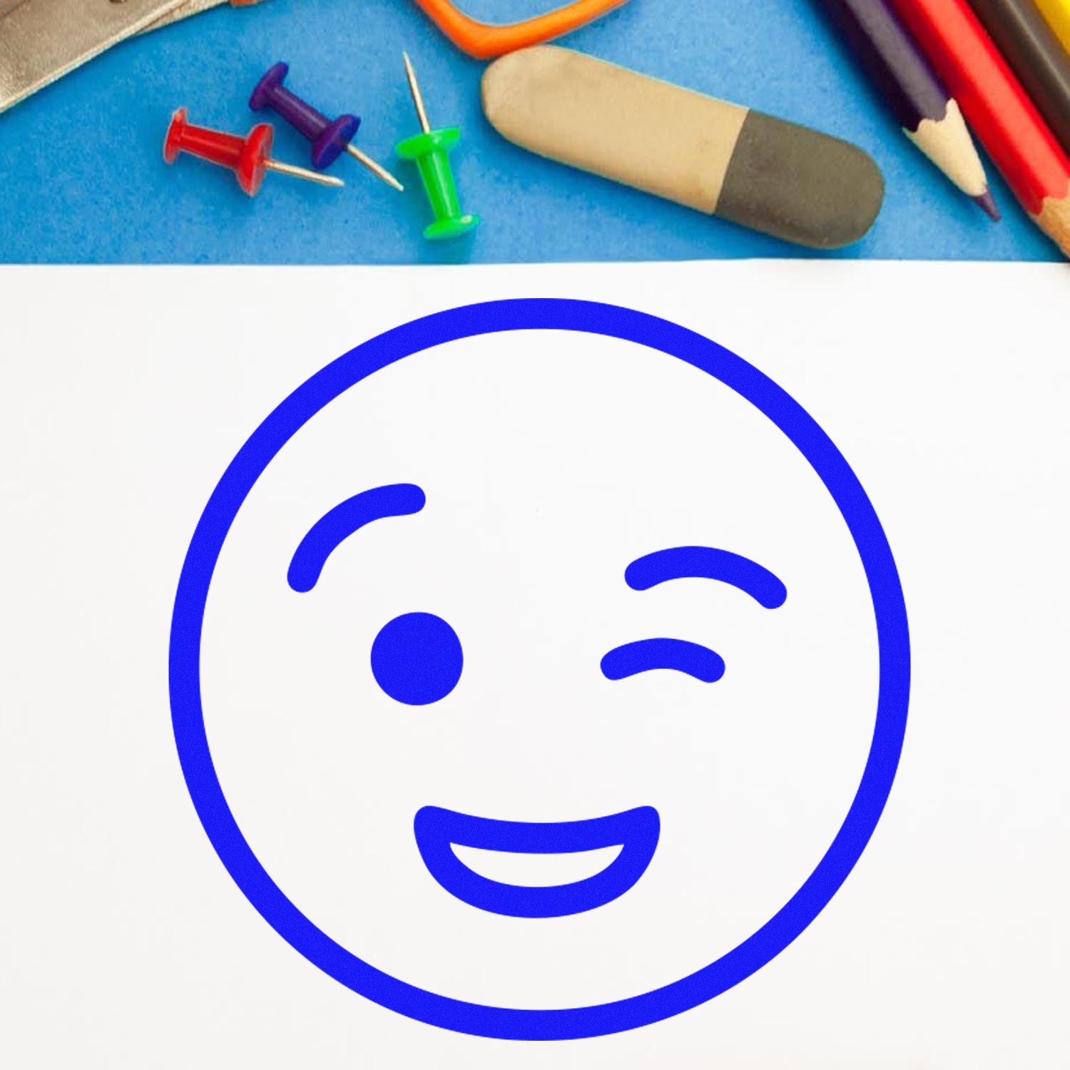 Self-Inking Cheeky Wink Emoticon Stamp on paper, featuring a playful winking face in blue. Surrounded by colorful push pins, pencils, and an eraser on a blue background.