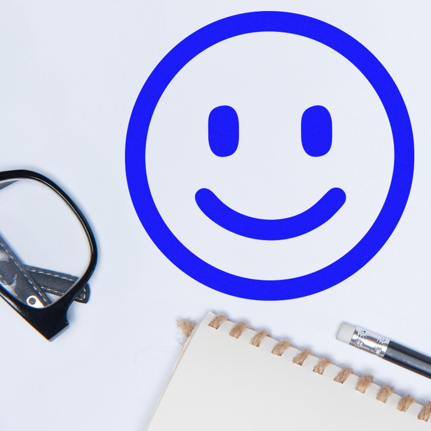 Self-Inking Happy Face Smiley Stamp in blue, shown on white paper with a notebook and glasses nearby. Perfect for adding cheerful marks to documents and crafts.