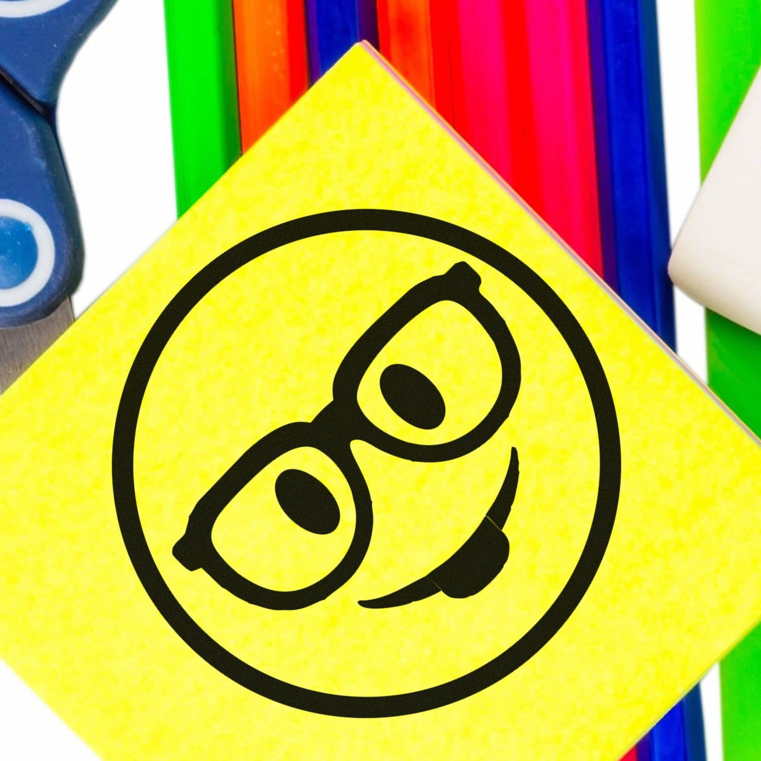 Self-Inking Smarty McGlasses Emotes Stamp on a yellow sticky note, featuring a playful smiley face with glasses. Surrounded by colorful pens and stationery for creative use.