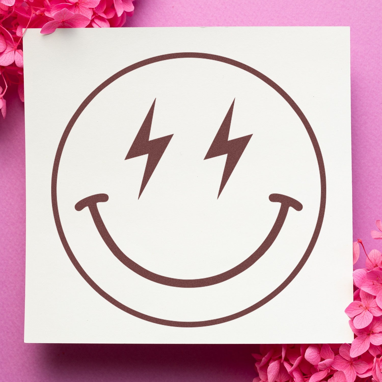 Wood Handle Electric Joy Emoji Rubber Stamp on pink background with flowers, featuring a smiling face with lightning bolt eyes. Perfect for crafting and personalizing projects.