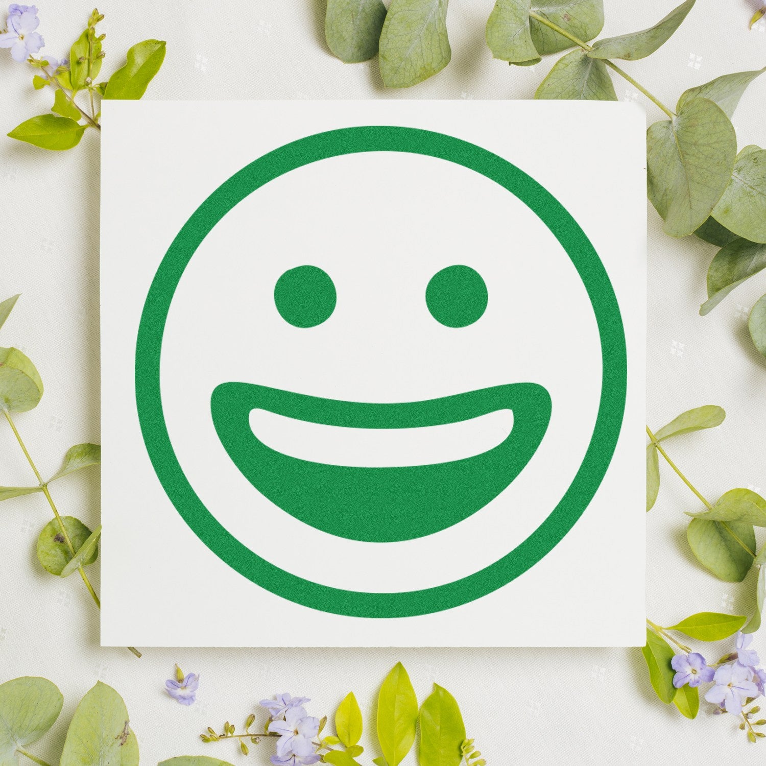 Self-Inking Classic Grin Emoji Stamp with a green smiling face design on a white background, surrounded by green leaves and small purple flowers. Perfect for adding fun to any project.