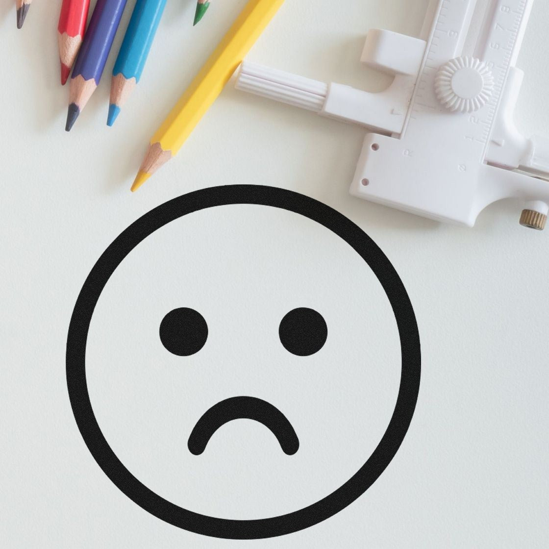 Self-Inking Sad Sam Emoticon Stamp on paper with colored pencils and a ruler, showcasing a clear, bold sad face design. Perfect for adding expressive touches to documents and crafts.