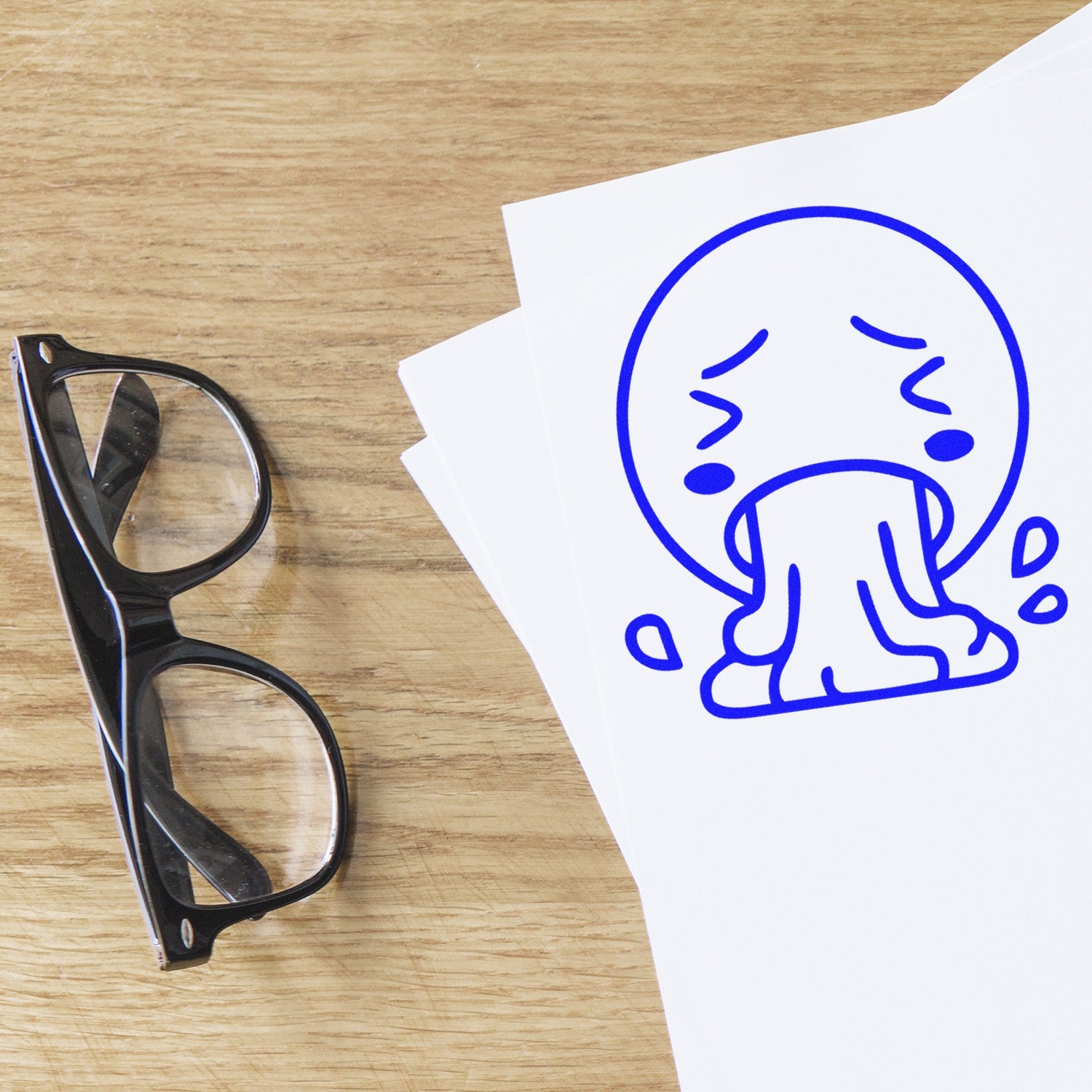 Self-Inking Queasy Quincy Pictogram Stamp on paper, featuring a blue cartoon face with a queasy expression, next to black glasses on a wooden surface.