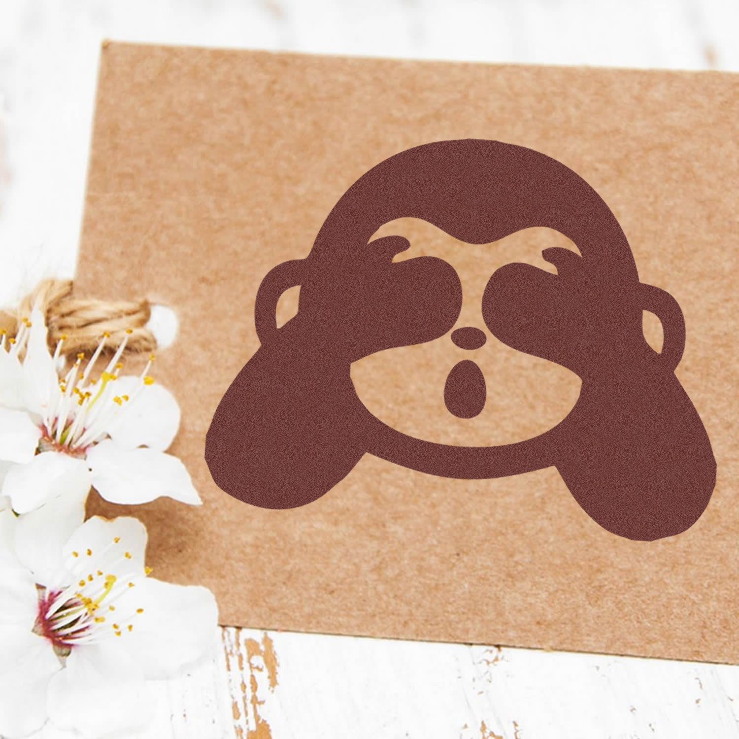 Wood Handle Blindfolded Monkey Emojicons Rubber Stamp on brown paper, featuring a playful monkey face with hands over eyes, next to white flowers on a rustic wooden surface.