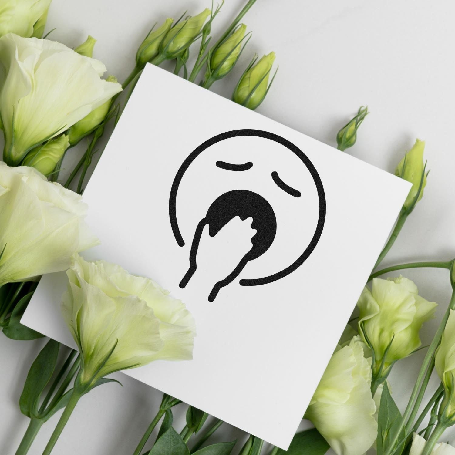 Self-Inking Yawning Yeti Emoji Stamp on white paper surrounded by green and white flowers, showcasing a yawning face design. Perfect for adding a playful touch to stationery.