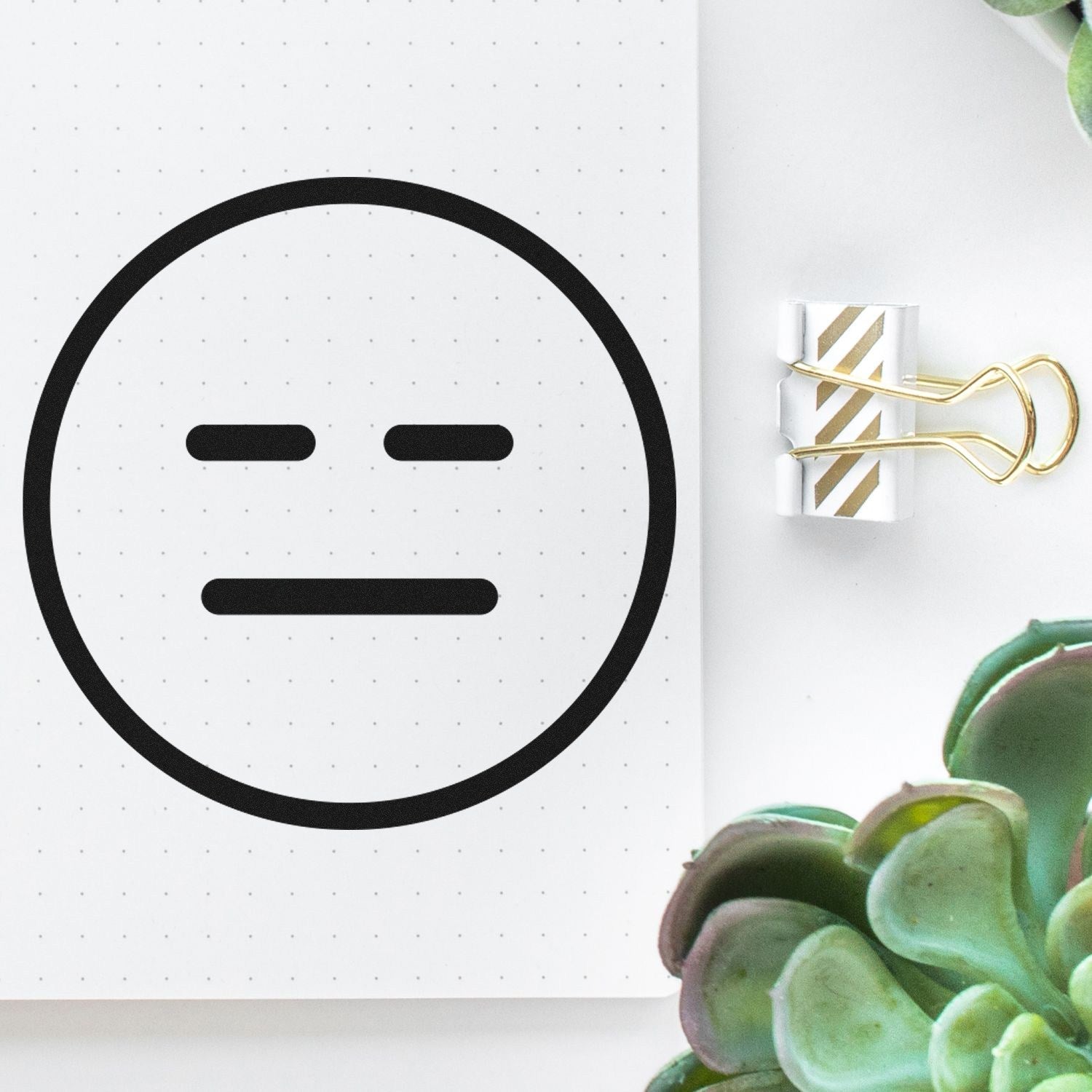 Wood Handle Expressionless Eddie Emoji Rubber Stamp on dotted paper, featuring a simple expressionless face design, with a gold-striped binder clip and green succulents nearby.