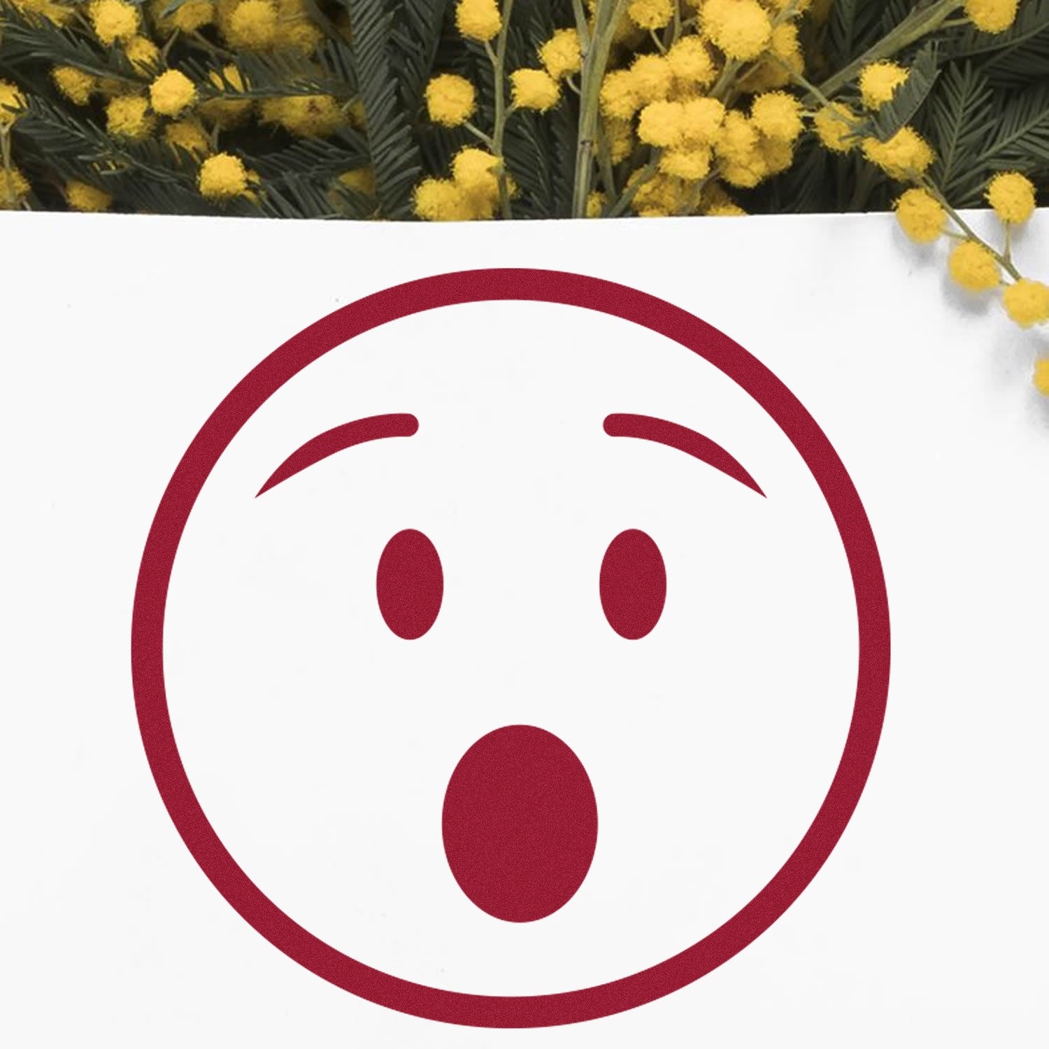 Self-Inking Amazed Andy Emotes Stamp with a surprised face design, shown on white paper against a background of yellow flowers. Perfect for adding expressive touches to crafts and documents.