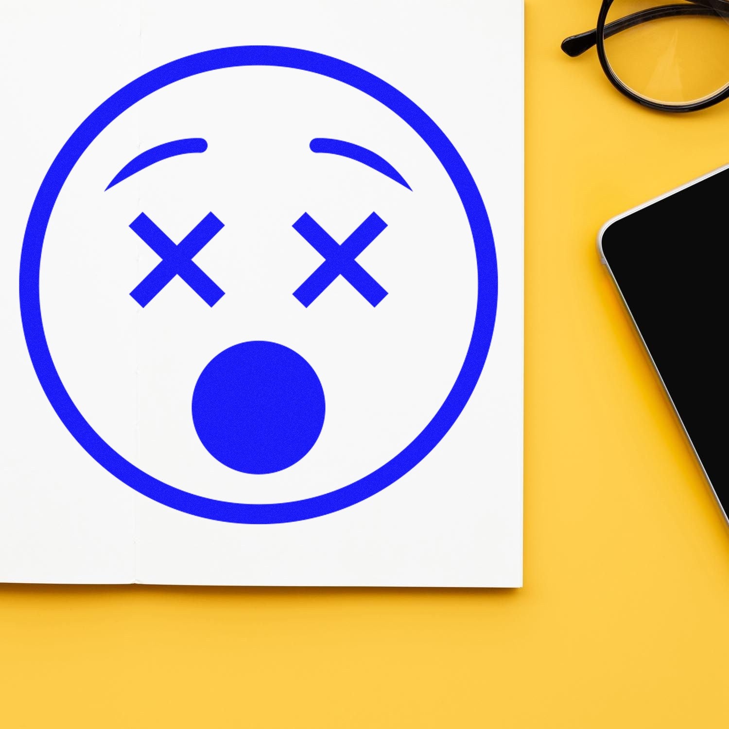 Image of the Self-Inking X'd Out Facial Expressions Stamp, featuring a blue, crossed-out eyes and open mouth design on white paper, with a yellow background and nearby glasses and smartphone.