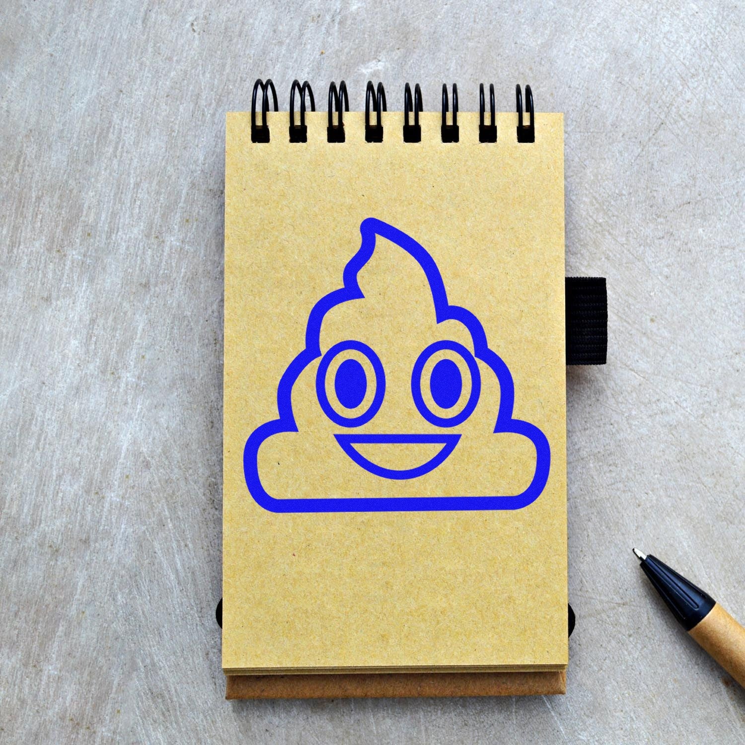 Wood Handle Smiling Poop Emojicons Rubber Stamp on a notebook, featuring a blue smiling poop emoji design. Perfect for adding a playful touch to stationery and crafts.