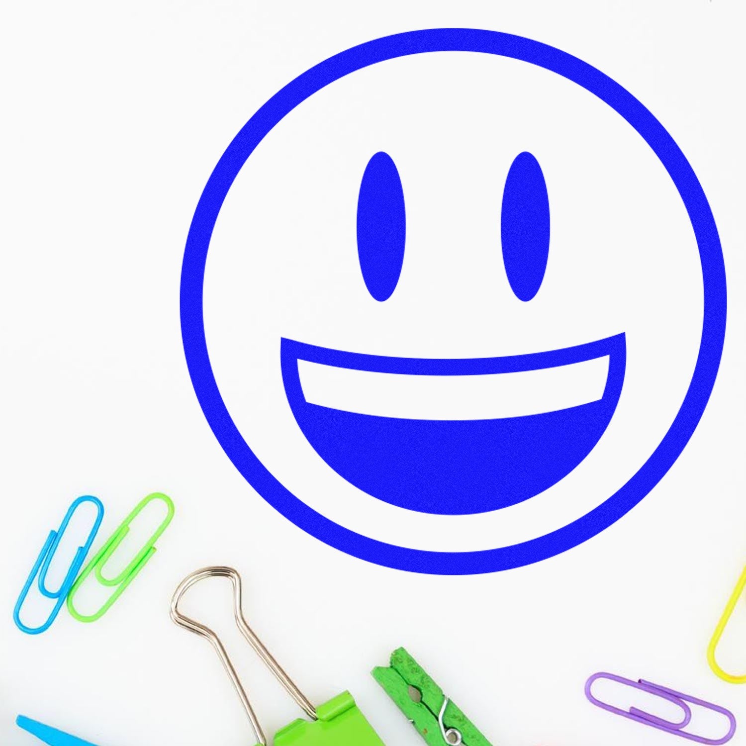 Self-Inking Grinning Delight Emoji Stamp with a blue smiling face design, surrounded by colorful paper clips and a binder clip on a white background. Perfect for adding fun to your stationery.