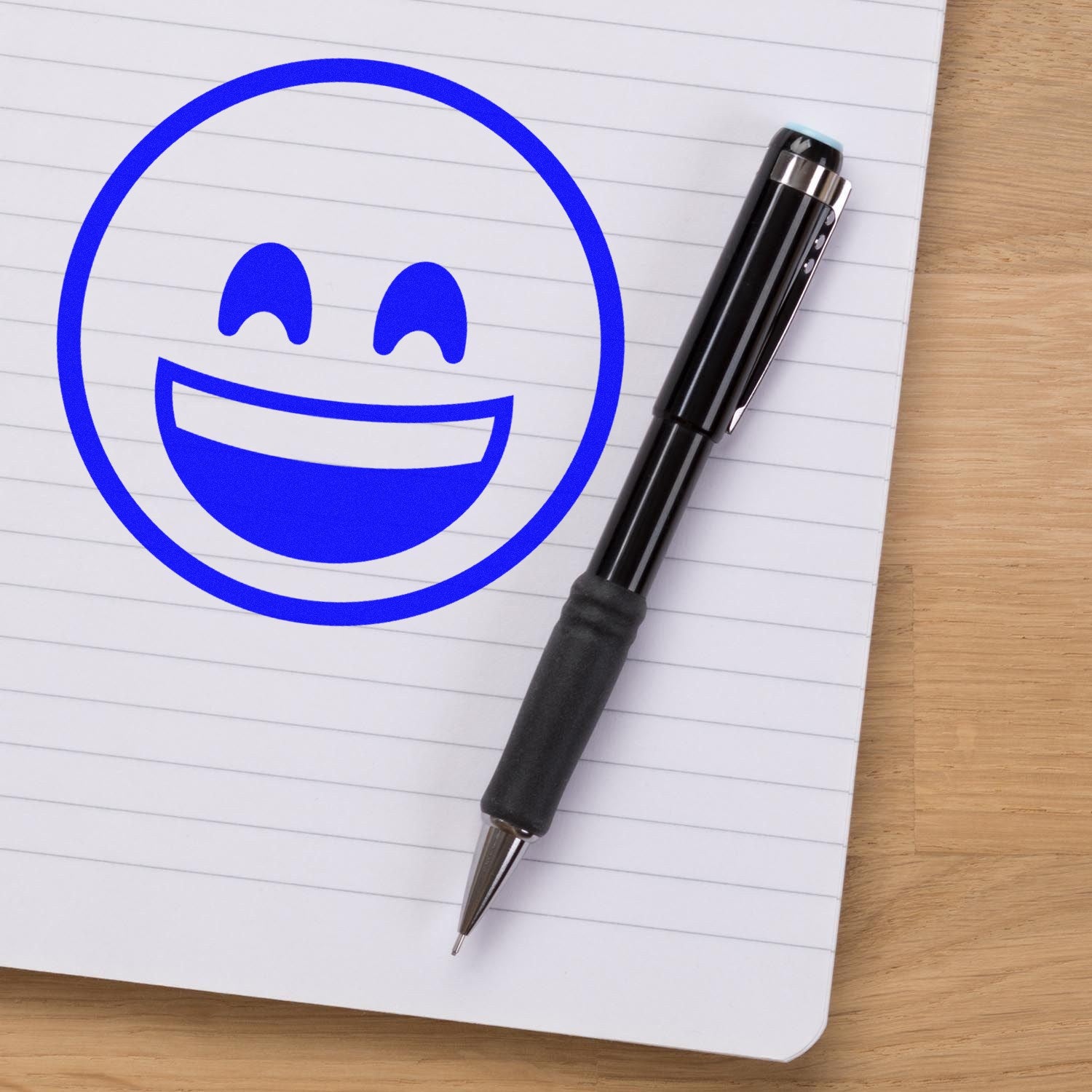 Wood Handle Ecstatic Eyes Emoji Rubber Stamp on lined paper, featuring a blue smiling emoji with closed eyes, next to a black pen on a wooden surface. Perfect for adding fun to notes.