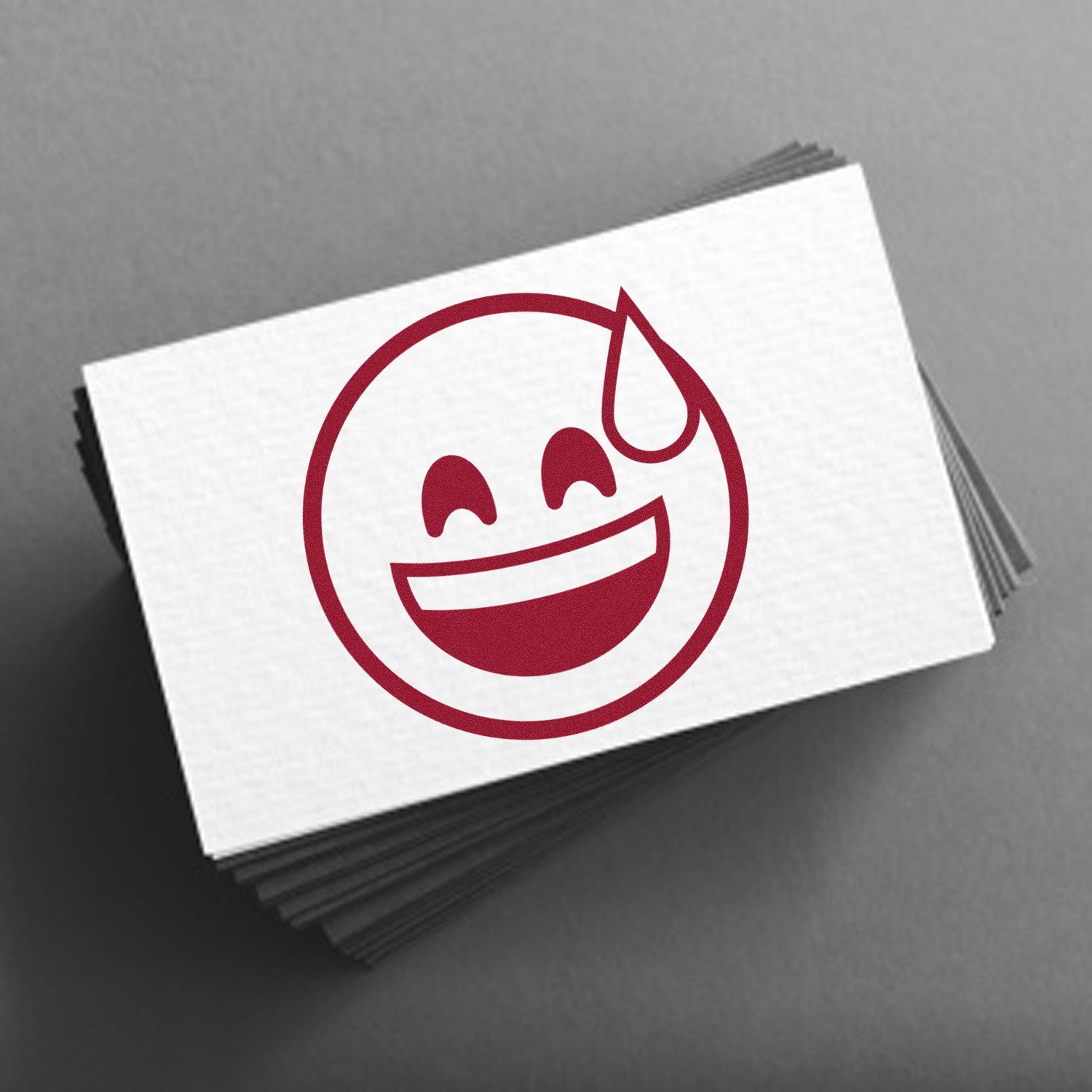 Image of the Self-Inking Blushing Chuckles Emoji Stamp, featuring a smiling face with a sweat drop, printed in red on a stack of white cards. Perfect for adding fun to your stationery.