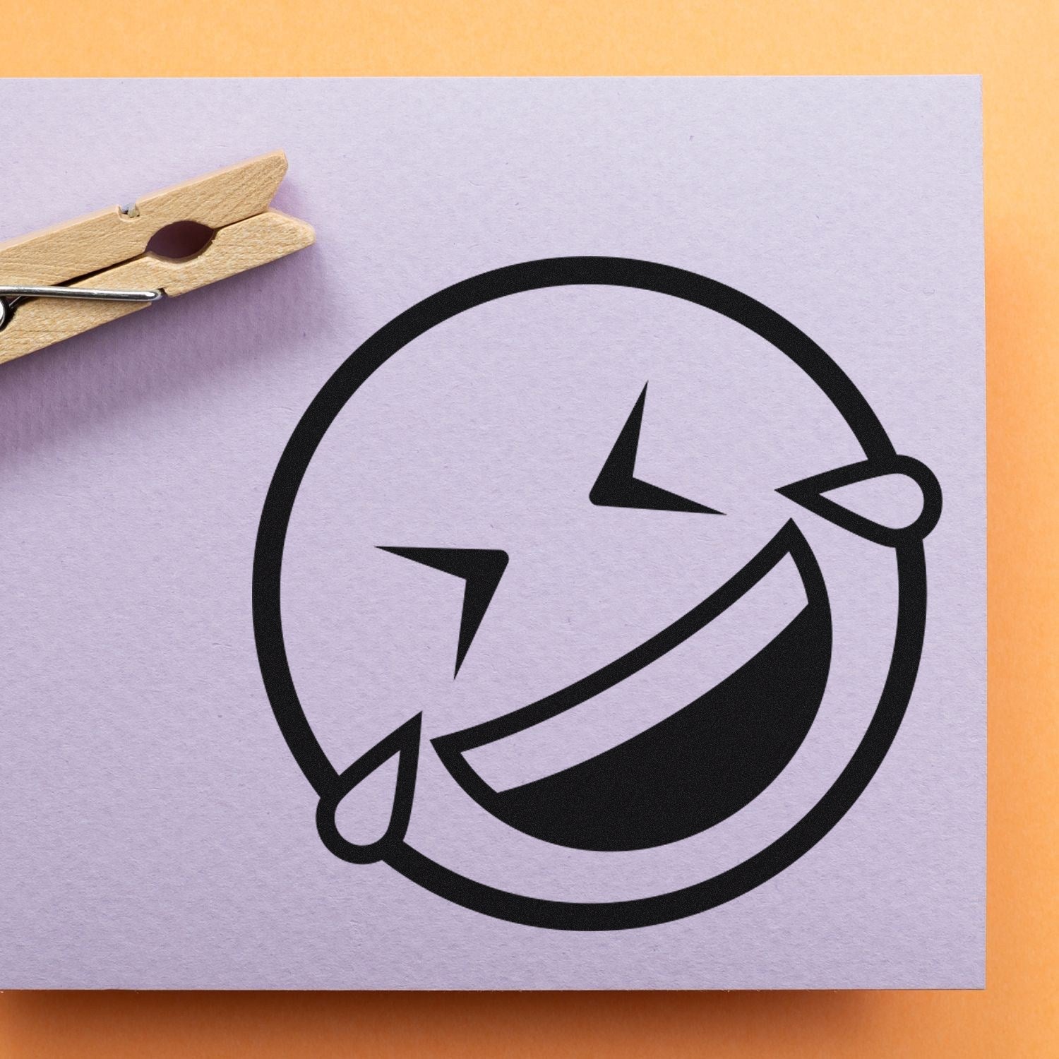 Wood Handle Laugh Riot Smiley Rubber Stamp imprint on paper, featuring a bold, laughing emoji design with tears of joy, set against a light background with a clothespin.