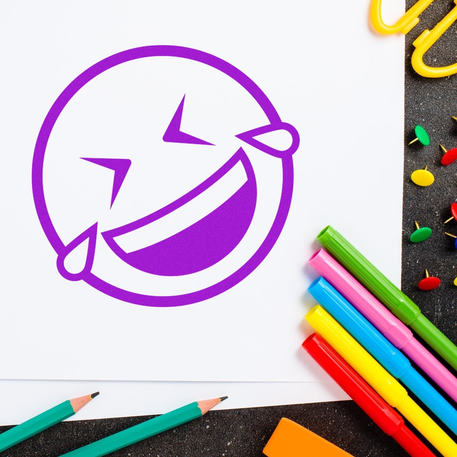 Self-Inking Laugh Riot Smiley Stamp in use, featuring a vibrant purple laughing emoji imprint on paper, surrounded by colorful pens and stationery. Perfect for adding fun to any project.