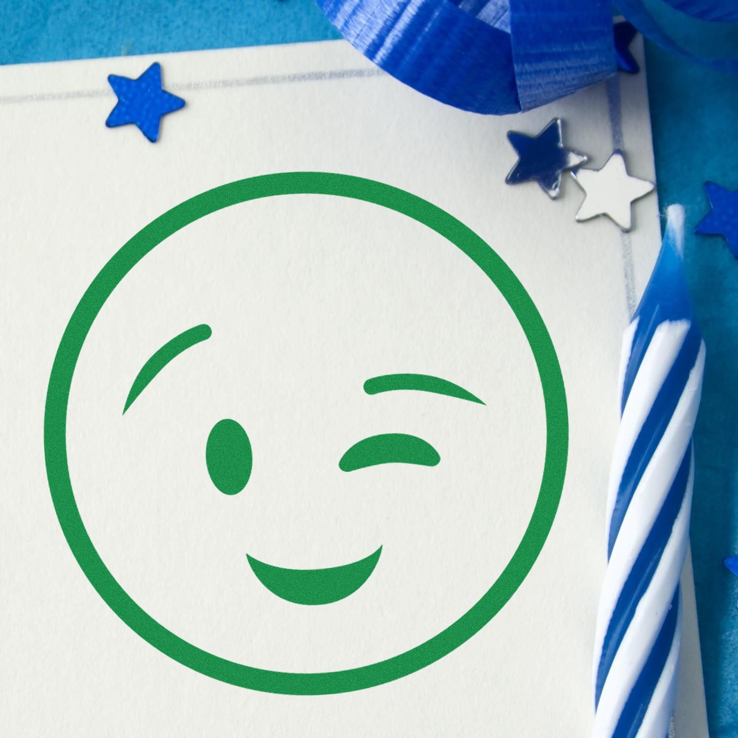 Wood Handle Winky Face Pictogram Rubber Stamp imprint on paper, featuring a playful green winky face. Surrounded by blue stars and a striped candle for a festive touch.