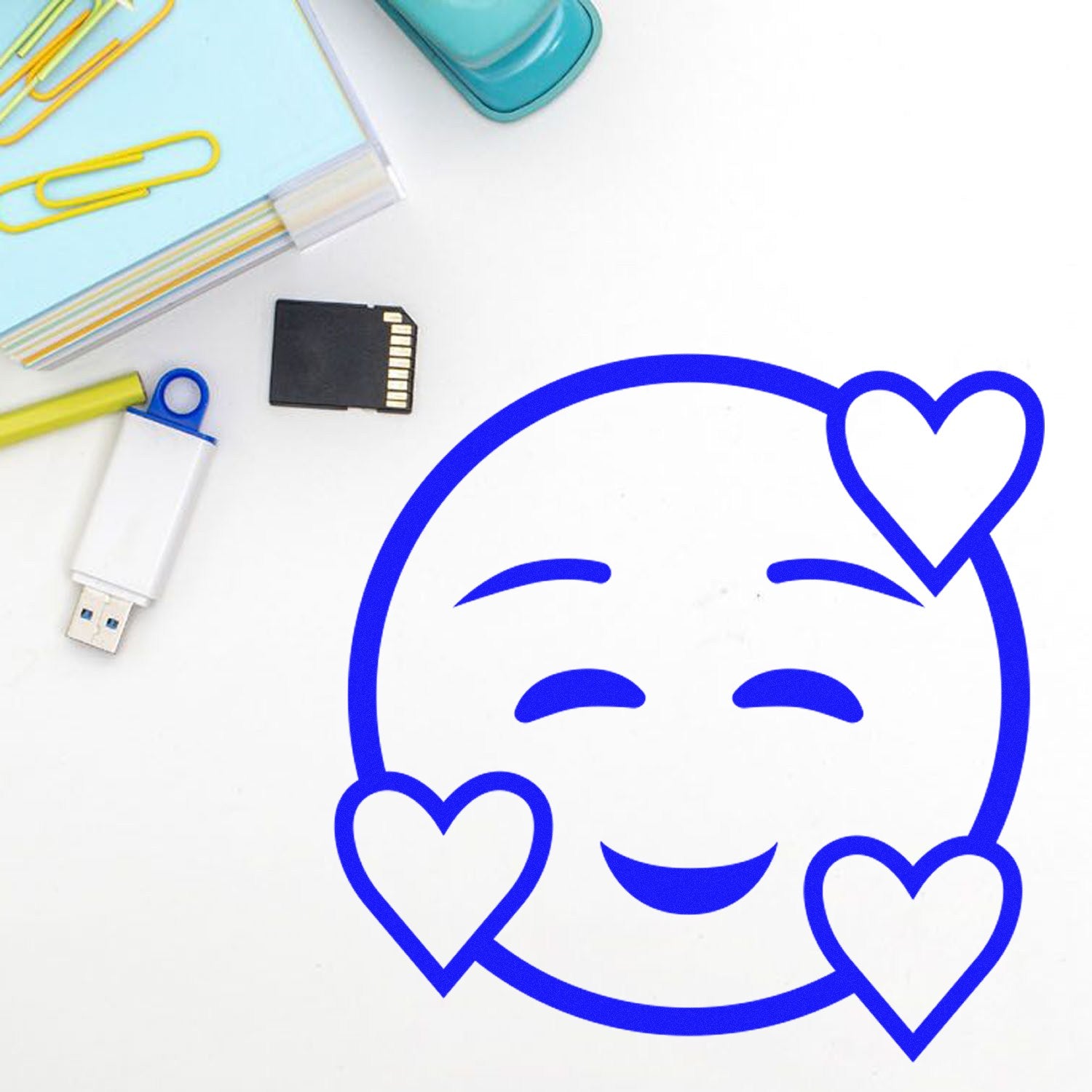 Self-Inking Love-Struck Emotes Stamp featuring a blue smiling face with heart eyes, surrounded by office supplies like a USB drive, SD card, and paper clips.