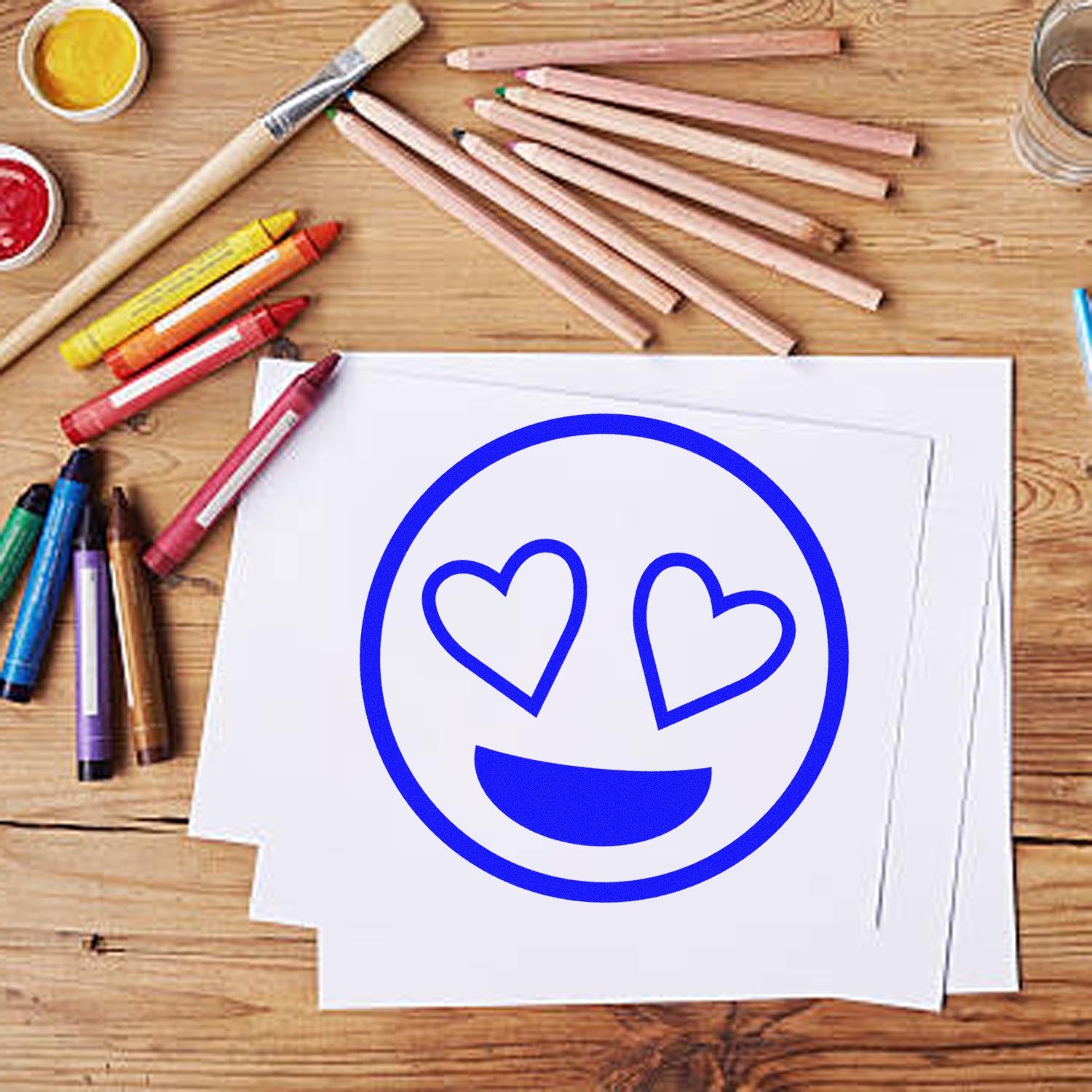 Wood Handle Heart-Eyes Reaction Rubber Stamp on a wooden table with art supplies, featuring a blue heart-eyes emoji design on white paper. Perfect for crafts and creative projects.