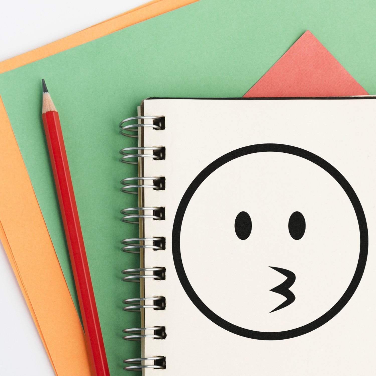 Image of the Self-Inking Whistling Wil Emojicons Stamp on a notebook, featuring a whistling face emoji design. The stamp is shown with colorful paper and a red pencil for context.