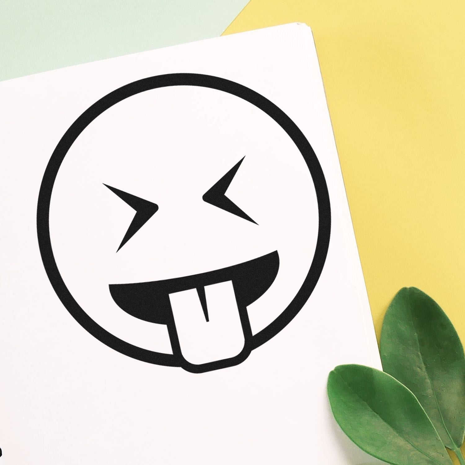 Wood Handle Crazy Face Emoji Rubber Stamp featuring a playful face with crossed eyes and a tongue out, perfect for adding fun to crafts. Set against a colorful background with green leaves.