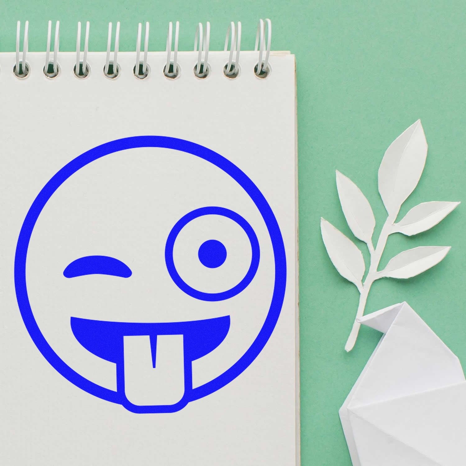 Wood Handle Goofy Wink Emoji Rubber Stamp on a notepad, featuring a playful blue winking face with tongue out. Perfect for adding fun to crafts and stationery.