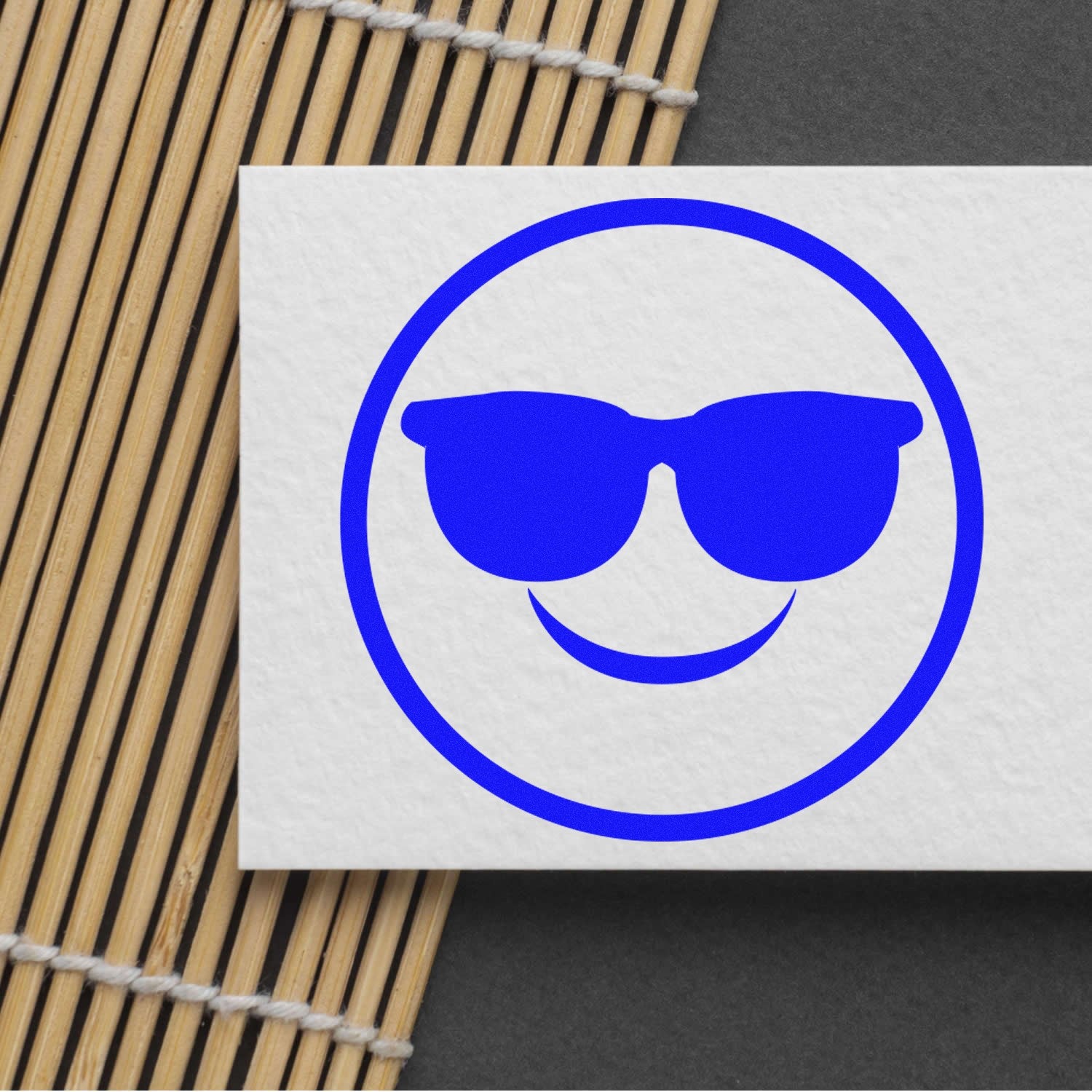 Wood Handle Cool Shades Pictogram Rubber Stamp featuring a blue smiley face with sunglasses on a white card, placed on a bamboo mat background.