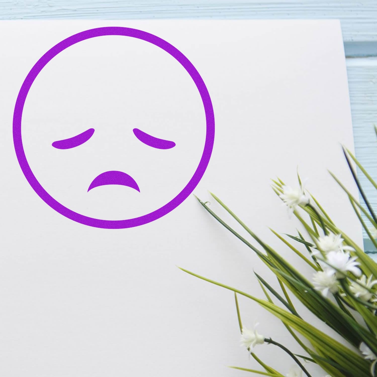 Self-Inking Sad Sammy Emojicons Stamp imprint on white paper, featuring a purple sad face emoji, next to green foliage on a light blue wooden surface.