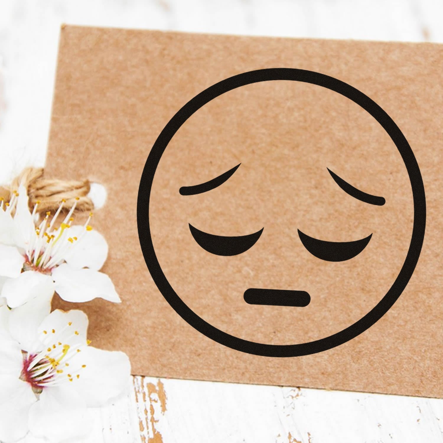 Wood Handle Silent Sorrow Emoji Rubber Stamp on brown paper, featuring a sad face design. Perfect for crafts and stationery. White flowers add a decorative touch.