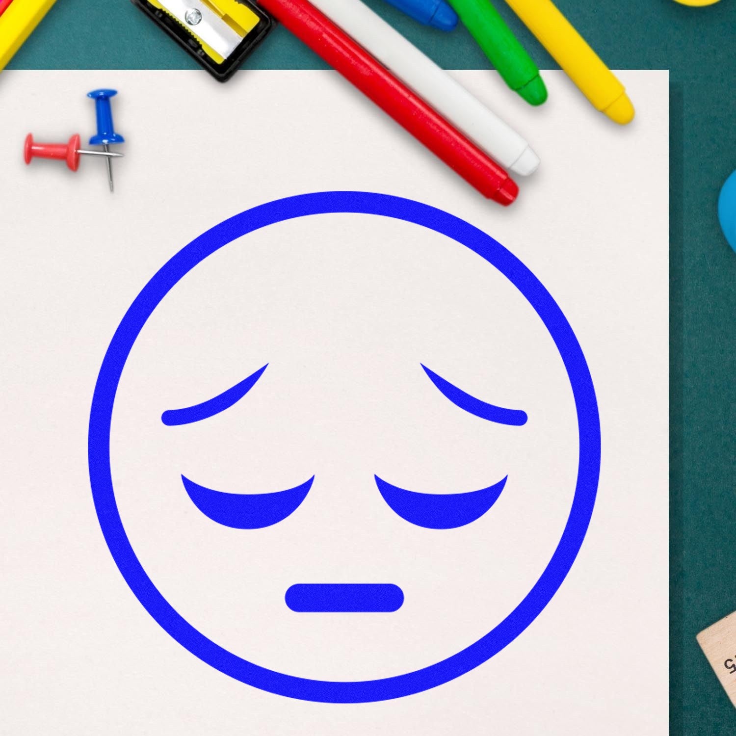 Wood Handle Silent Sorrow Emoji Rubber Stamp on paper, featuring a blue sad face. Surrounded by colorful pens and push pins, ideal for crafting and personalizing stationery.