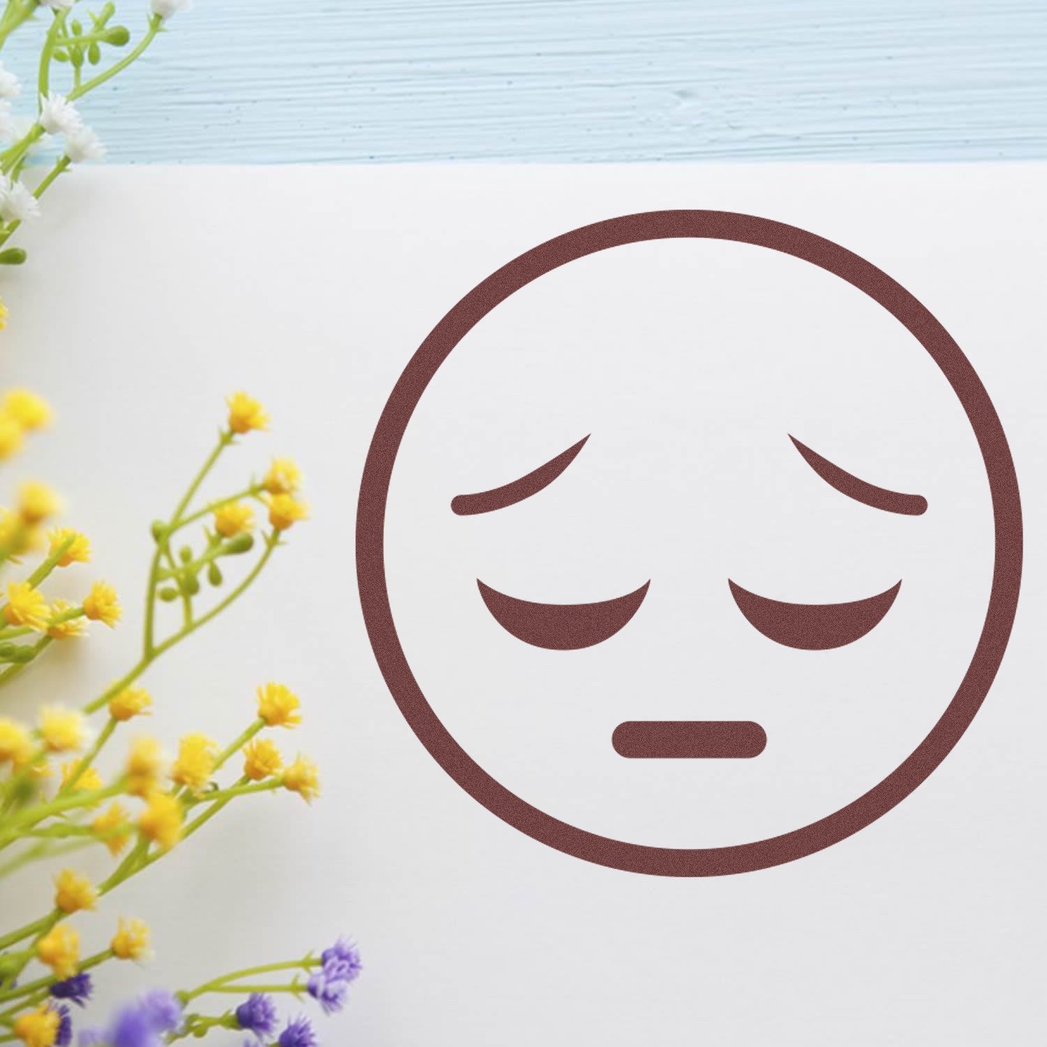 Wood Handle Silent Sorrow Emoji Rubber Stamp on paper, featuring a sad face design. Perfect for crafts and stationery. Surrounded by colorful flowers for a creative touch.