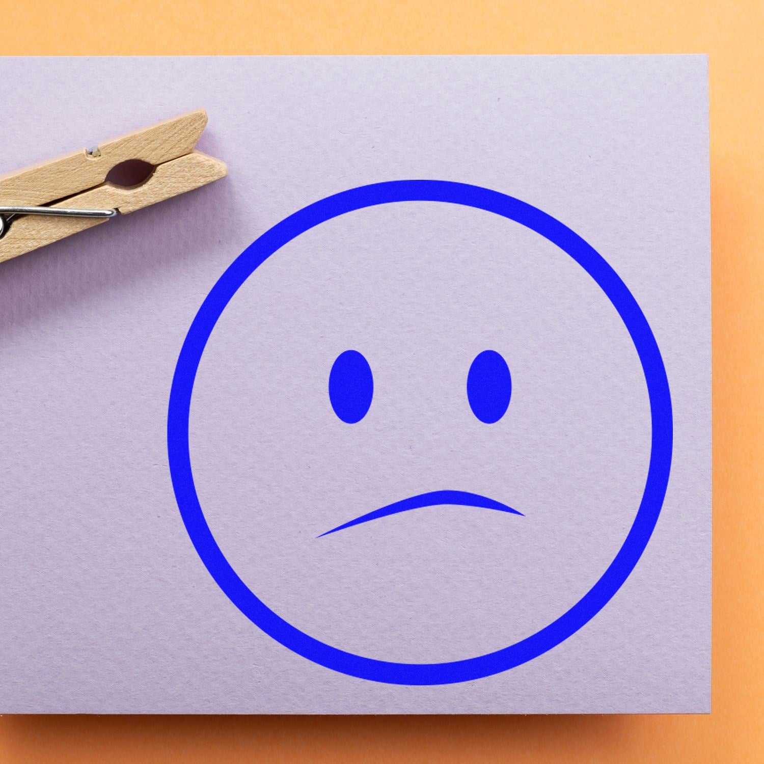 Wood Handle Solemn Simplicity Emoji Rubber Stamp creating a blue solemn face on paper, with a clothespin nearby. Perfect for crafting and personalizing stationery.