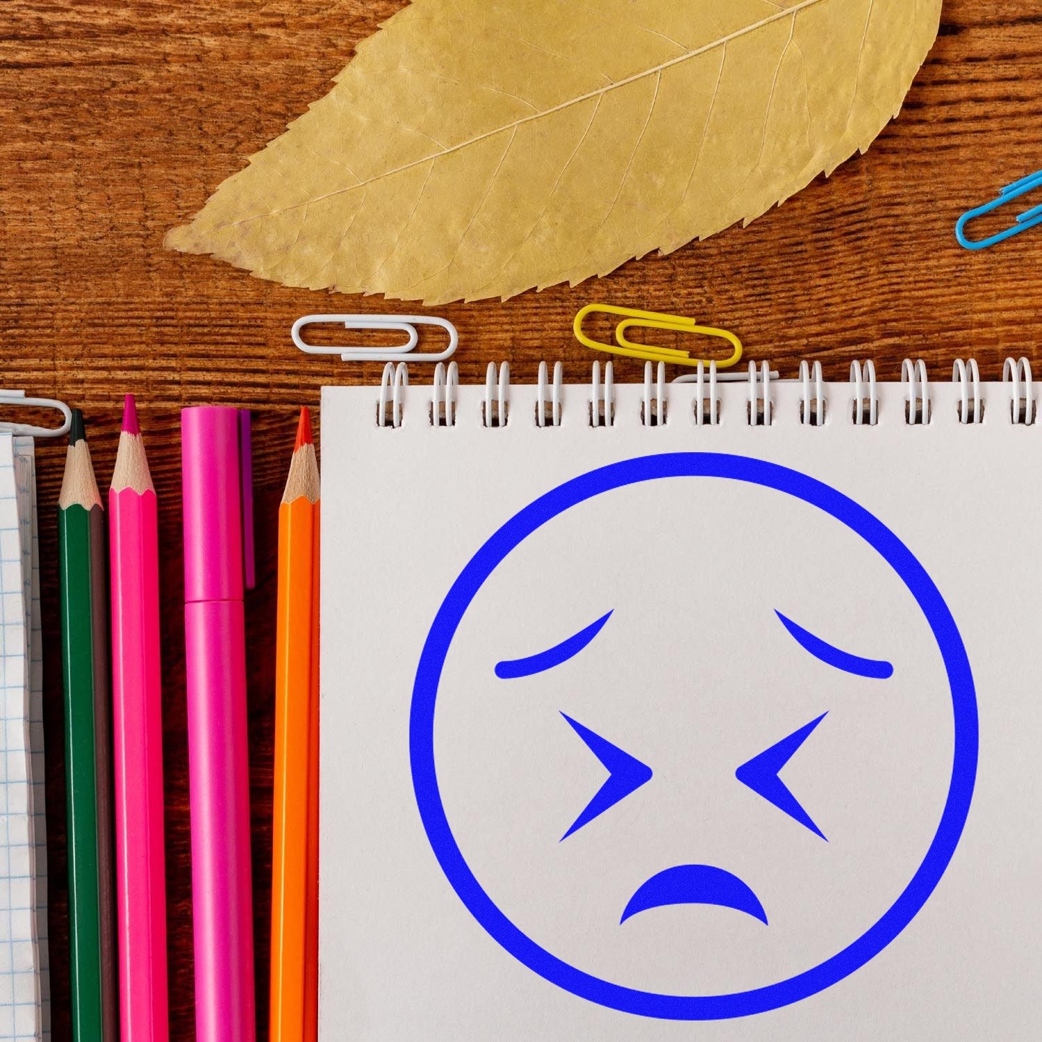 Self-Inking Starry Sadness Emoji Stamp on a notepad, surrounded by colorful pencils and paperclips, showcasing its blue sad face design. Perfect for adding a touch of emotion to your stationery.