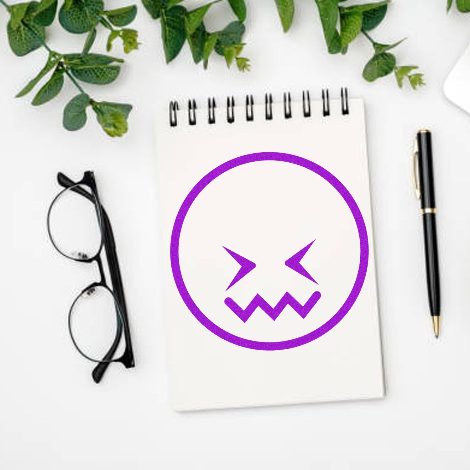 Wood Handle Zigzag Worry Emoji Rubber Stamp on a notepad, featuring a purple zigzag worry emoji. Glasses and a pen are placed nearby, with green leaves in the background.