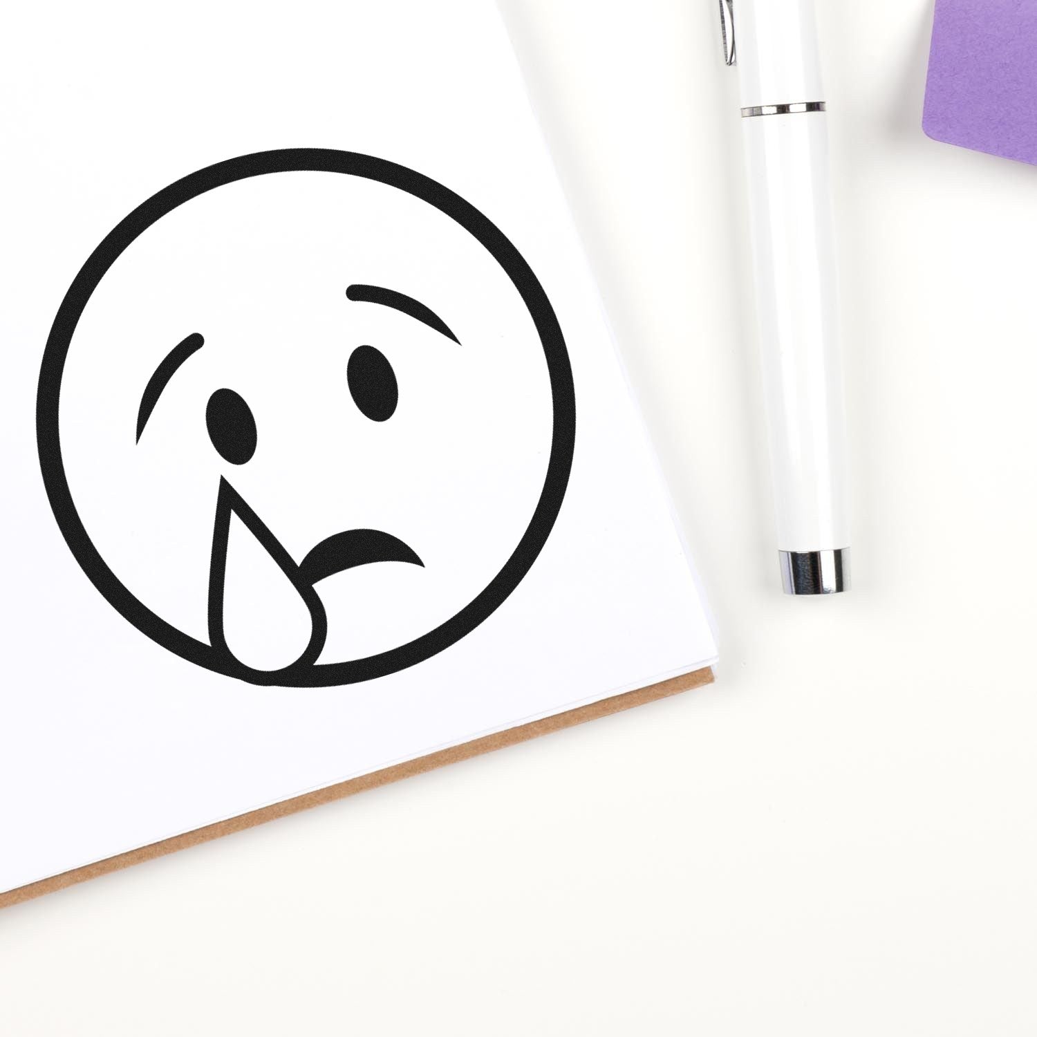 Wood Handle Teary Timmy Emoji Rubber Stamp imprint on paper, showing a crying face emoji. Includes a white pen and purple notepad in the background.