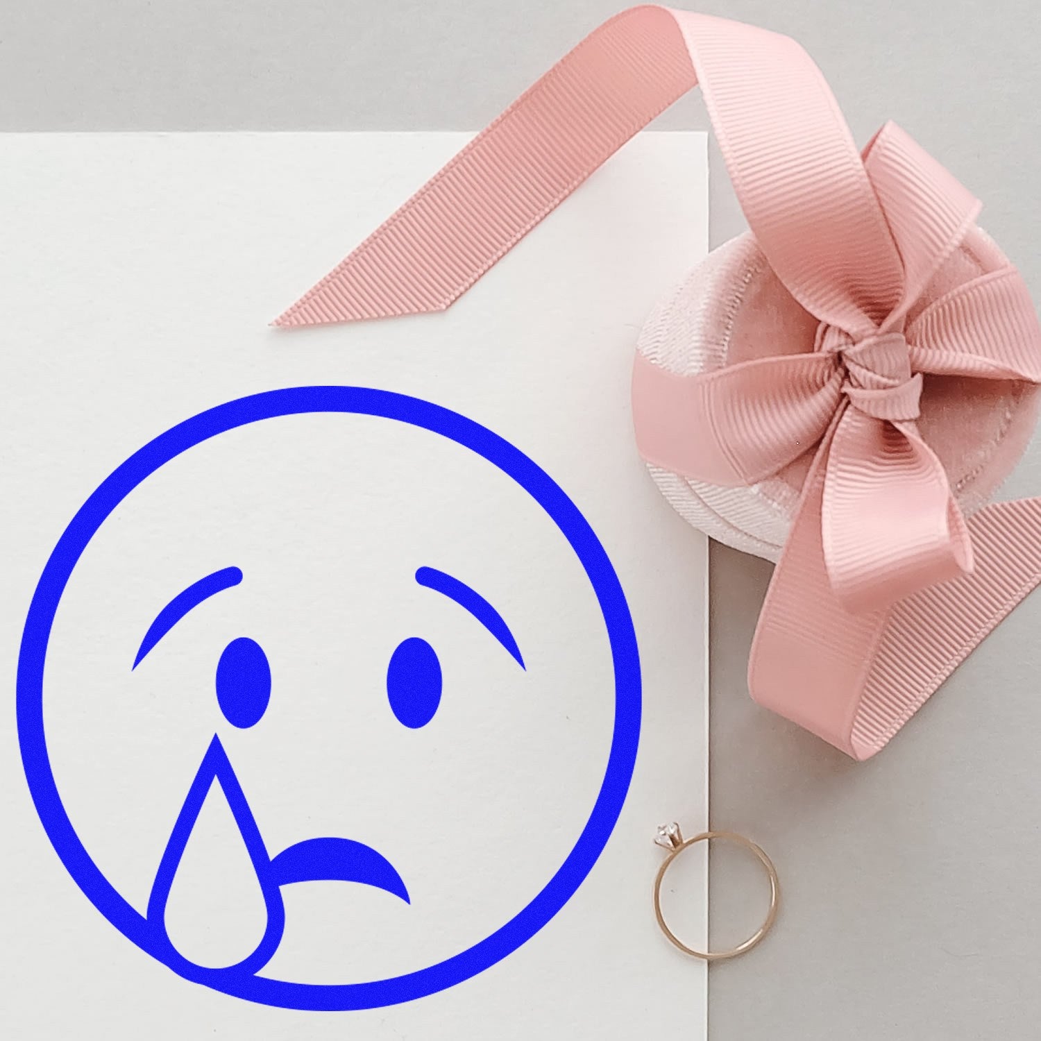 Self-Inking Teary Timmy Emoji Stamp creating a blue teary face on white paper, next to a pink ribbon and a ring. Perfect for adding a playful touch to crafts and letters.