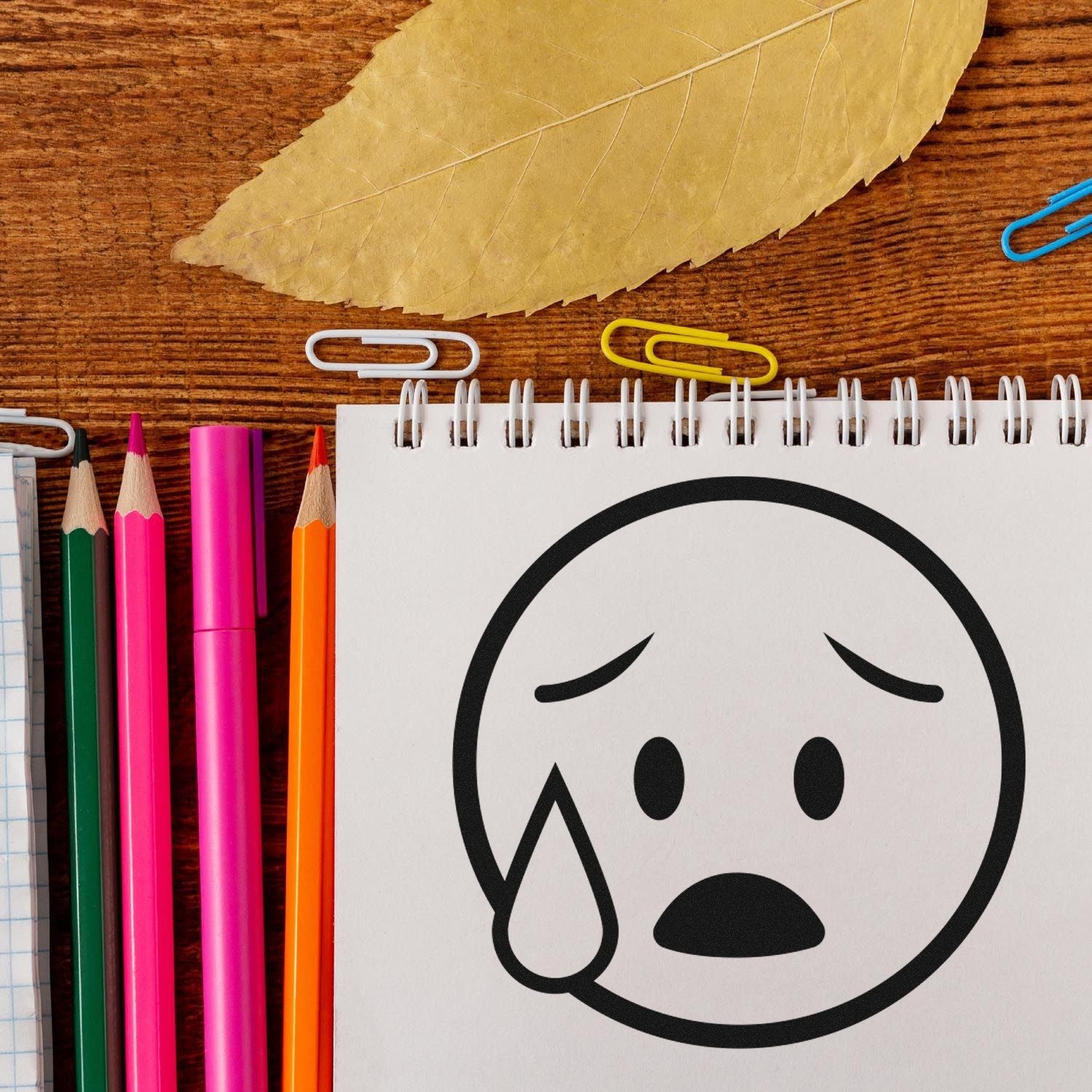 Self-Inking Tearful Timmy Facial Expressions Stamp on notepad, surrounded by colorful pencils and paperclips, showcasing a crying face design. Perfect for adding emotion to your stationery.