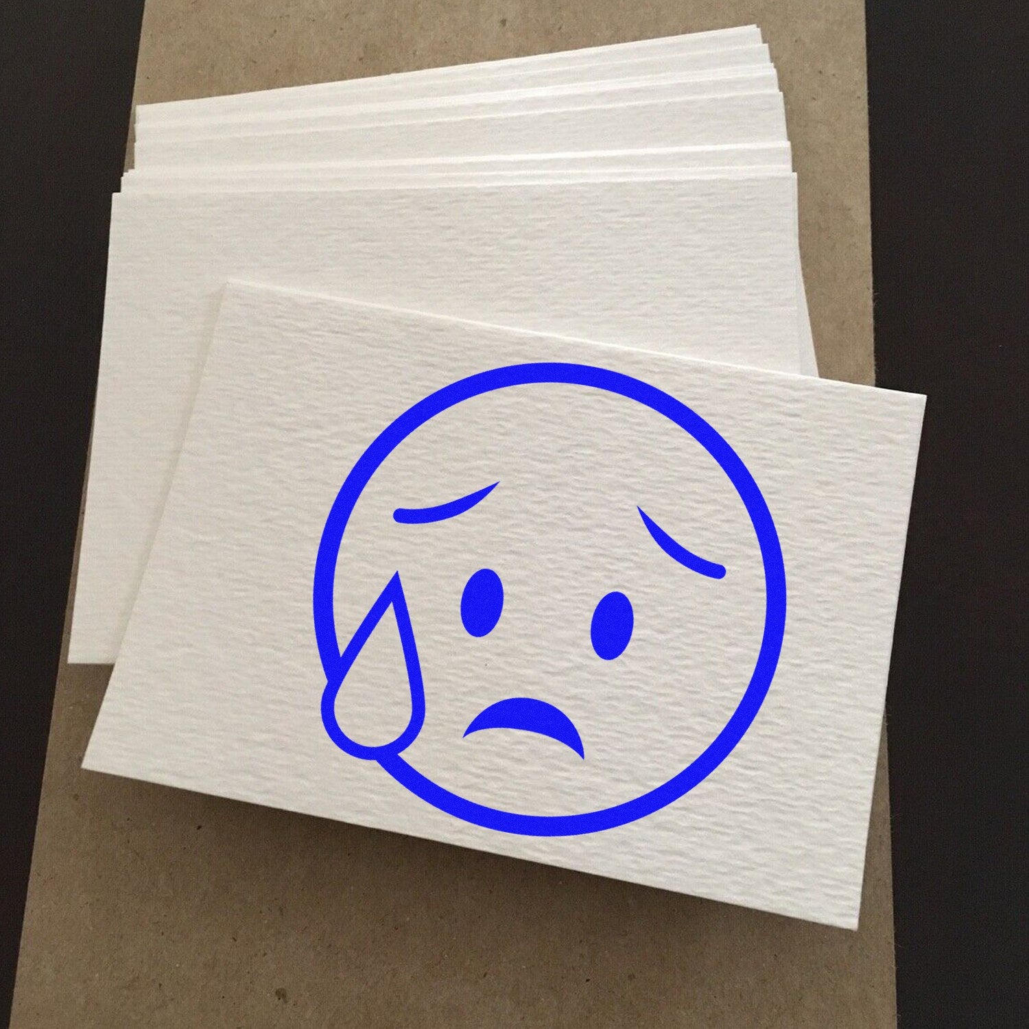 Wood Handle Weepy Wanda Emojicons Rubber Stamp featuring a blue sad face emoji with a tear, displayed on textured white paper sheets.