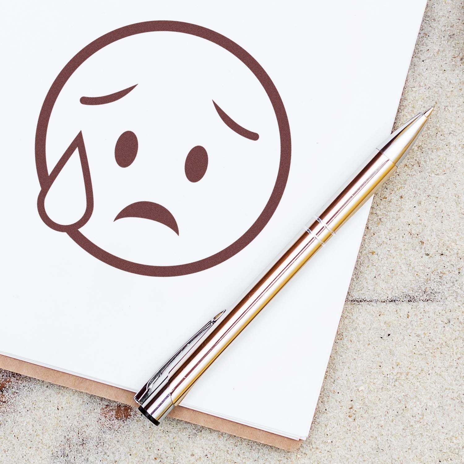 Self-Inking Weepy Wanda Emojicons Stamp on paper, featuring a sad emoji with a teardrop. Includes a sleek pen beside the stamped image, showcasing the product's design and functionality.