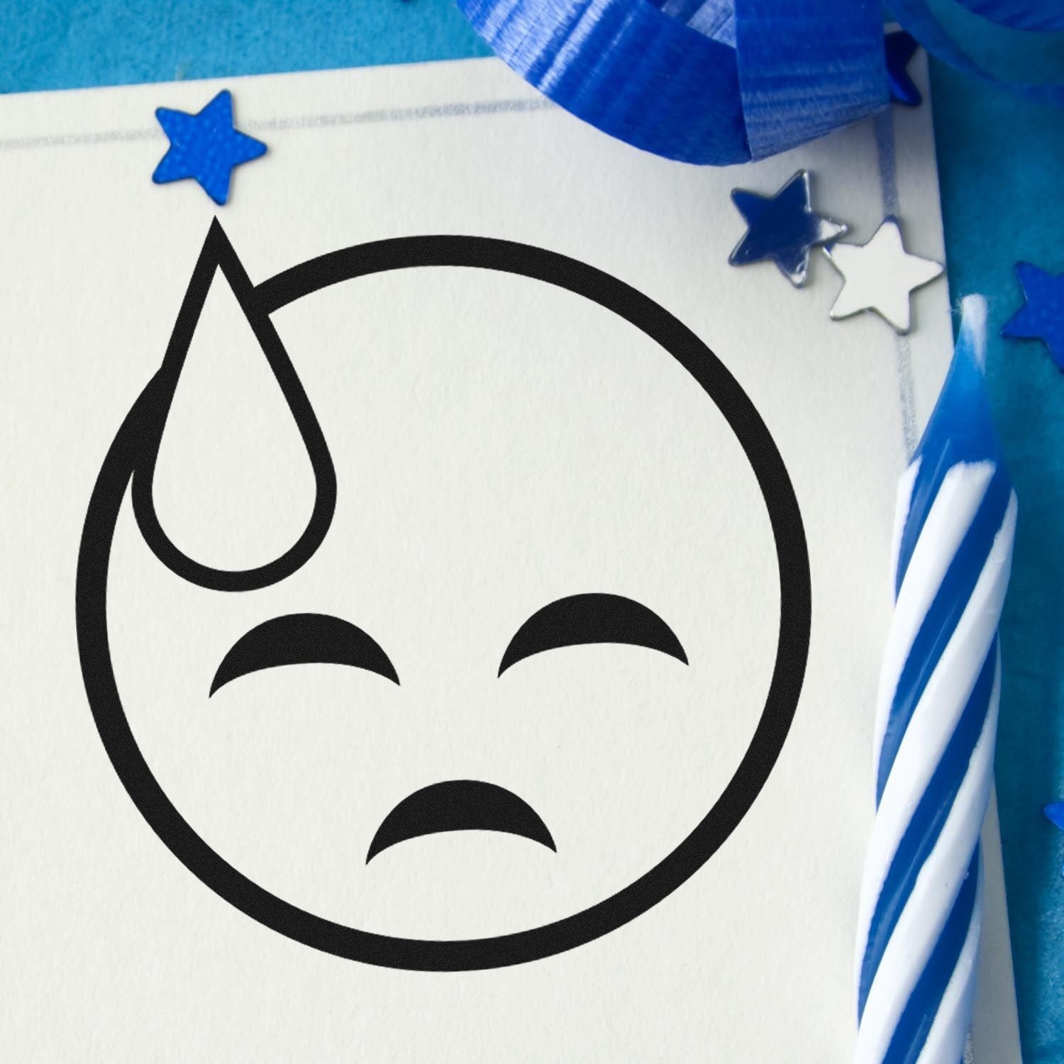 Wood Handle Sweaty Sadface Emoji Rubber Stamp on white paper, featuring a simple sad face with a sweat drop. Blue and white candle and star decorations in the background.