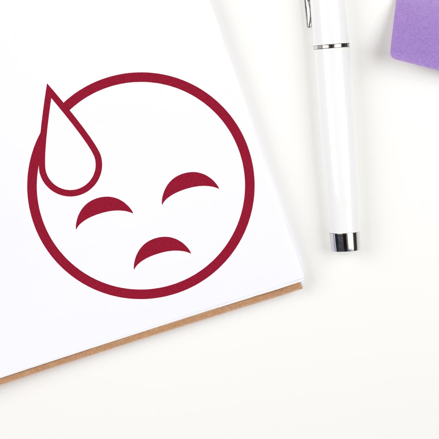 Wood Handle Sweaty Sadface Emoji Rubber Stamp imprint on paper, featuring a red sad face with a sweat drop. Includes a pen and purple notepad in the background.