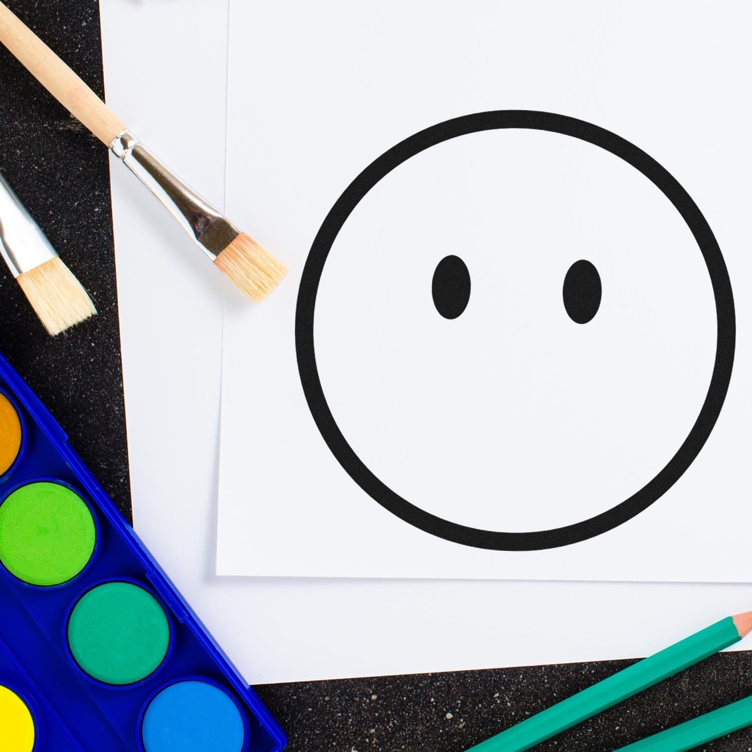 Self-Inking Speechless Sam Emoji Stamp on paper, surrounded by paintbrushes and watercolor palette. Perfect for adding fun, expressive touches to crafts and stationery.