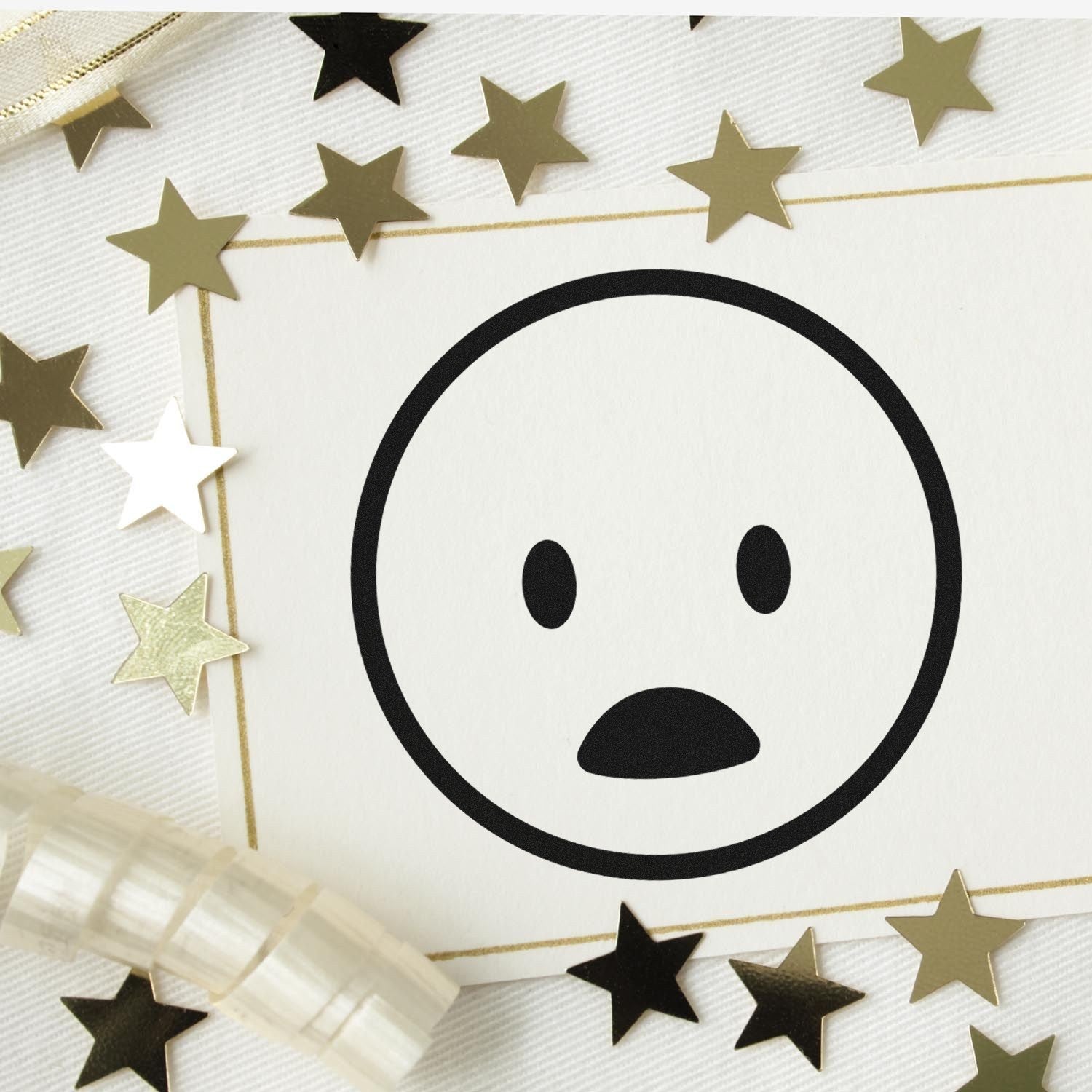 Self-Inking Surprised Smiley Emoji Stamp on white paper surrounded by gold stars and ribbon, perfect for adding fun expressions to crafts and stationery.