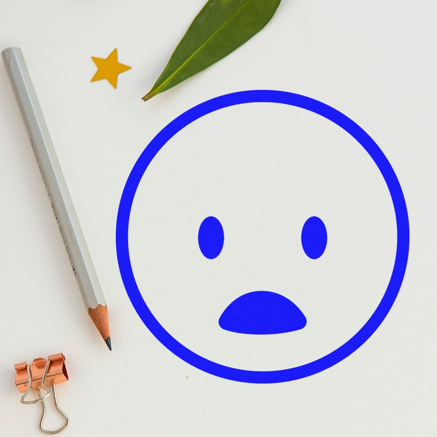 Wood Handle Surprised Smiley Emoji Rubber Stamp on paper, featuring a blue surprised face. Includes a pencil, leaf, star sticker, and copper binder clip for a creative workspace vibe.