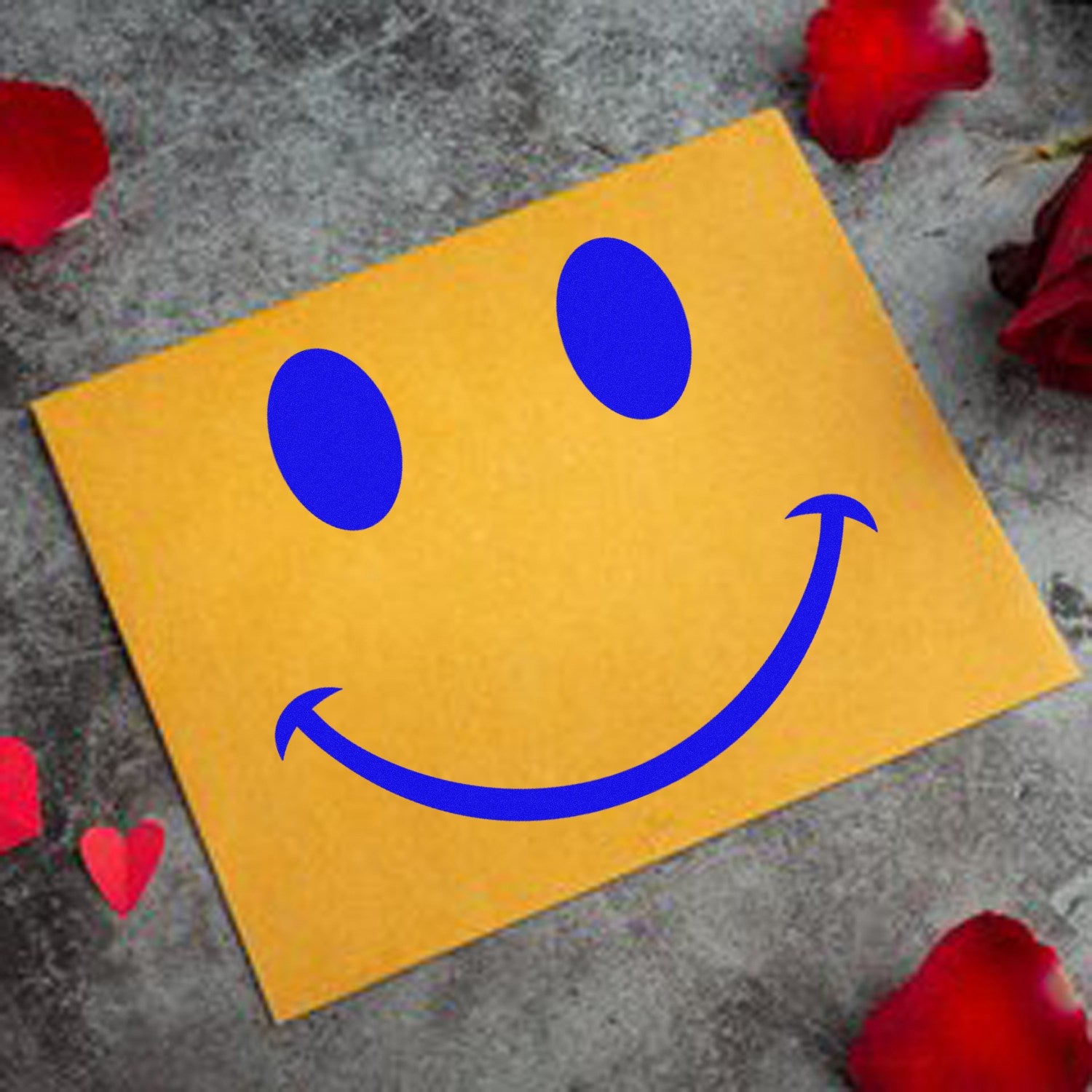 Wood Handle Cheeky Smirk Emoji Rubber Stamp on a gray surface with red rose petals, featuring a playful blue smirk emoji design on a yellow card.