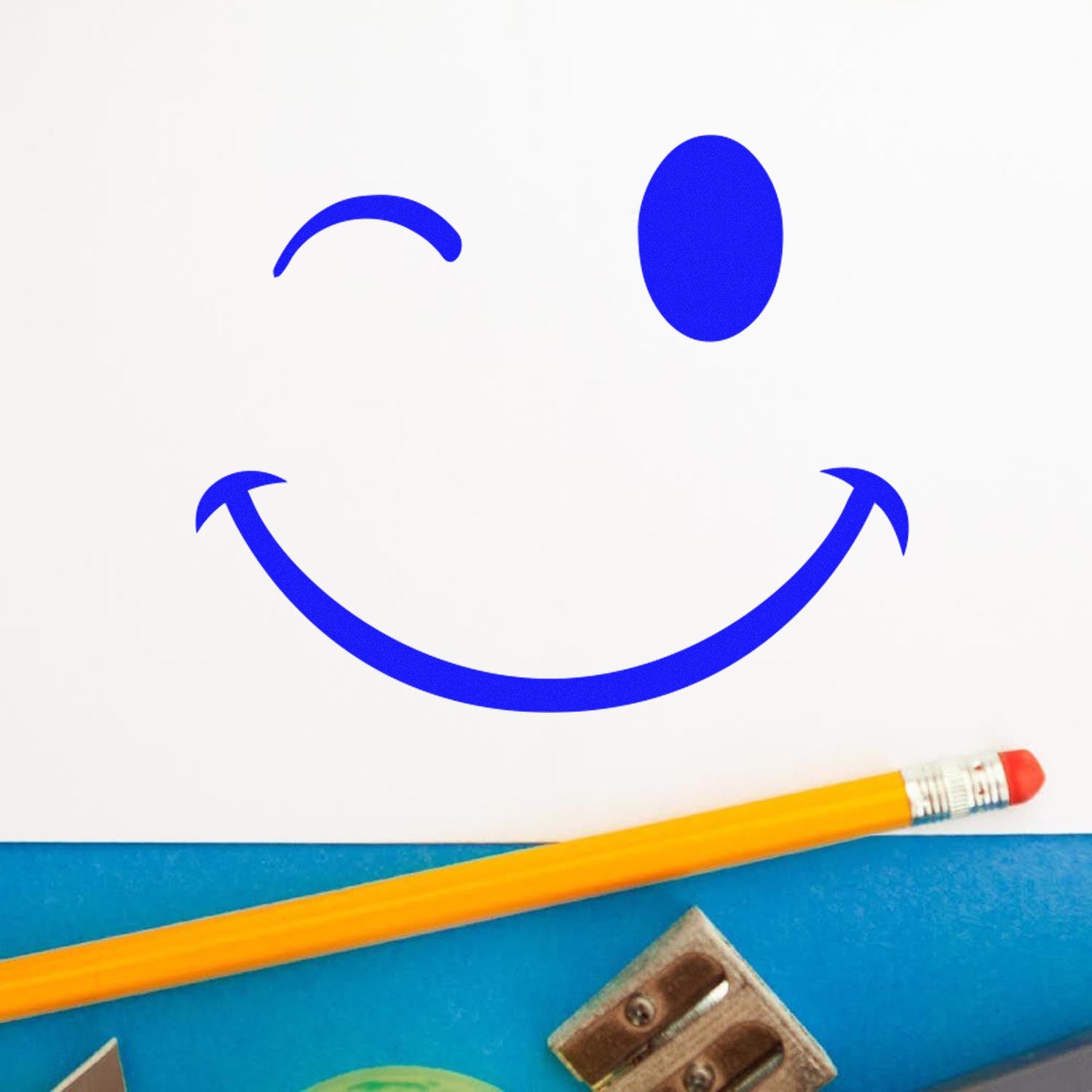 Image of a Self-Inking Winky Smirk Emoji Stamp imprint in blue on white paper, with a pencil and sharpener nearby. Perfect for adding a playful touch to notes and crafts.