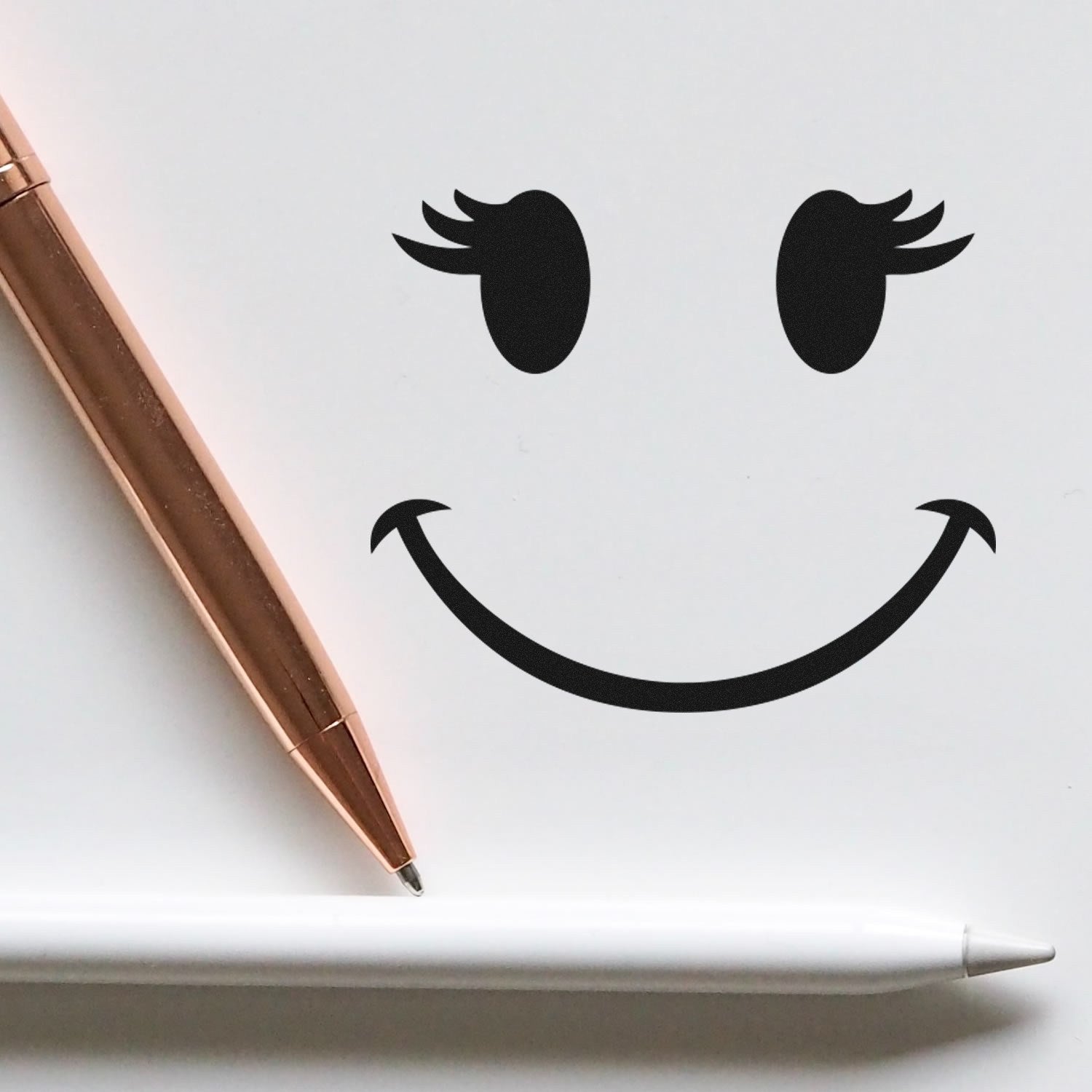 Image of the Wood Handle Fluttery Lashes Emoji Rubber Stamp creating a smiling face with fluttery lashes on white paper, alongside a rose gold pen and a white stylus.