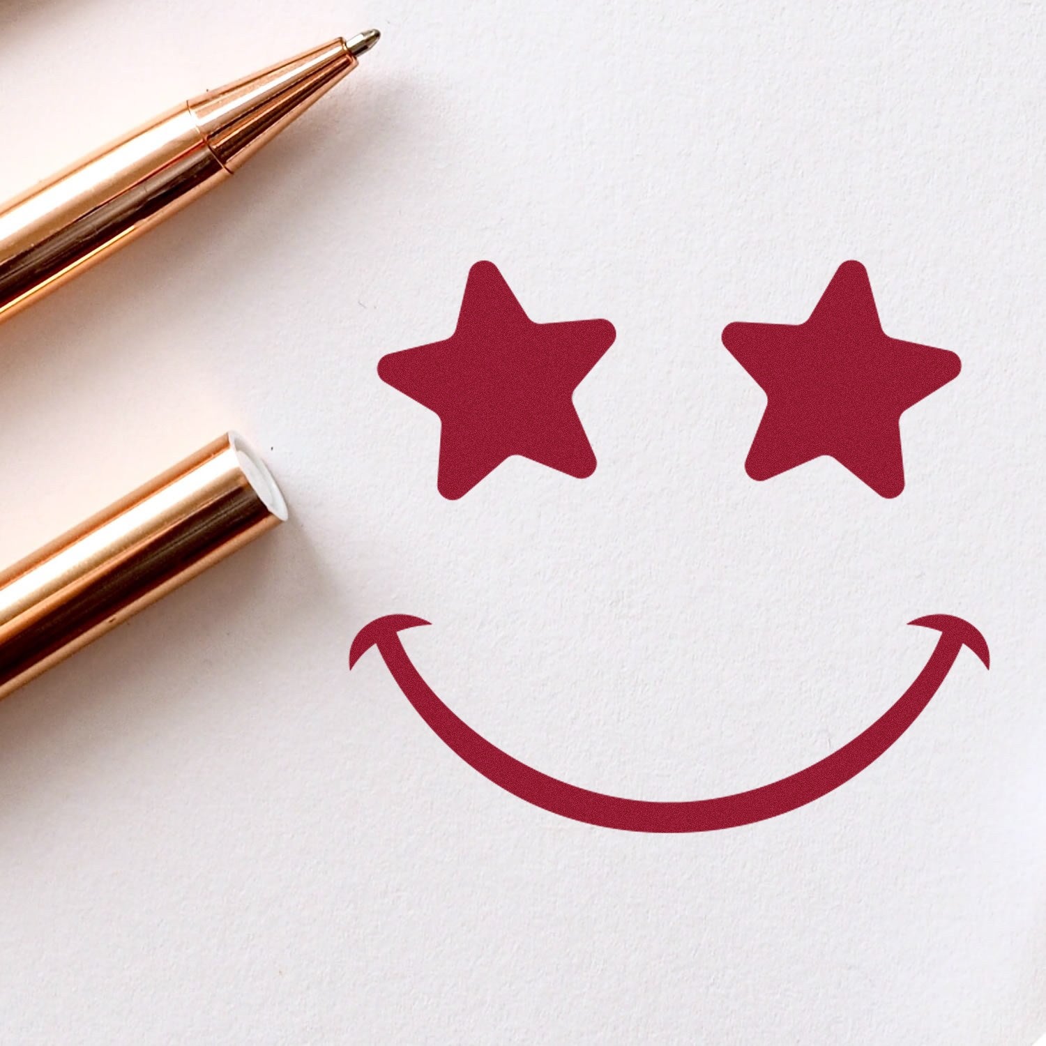 Self-Inking Starstruck Smile Smiley Stamp with red star eyes and curved smile on white paper, next to a rose gold pen. Perfect for adding fun, expressive designs to your projects.