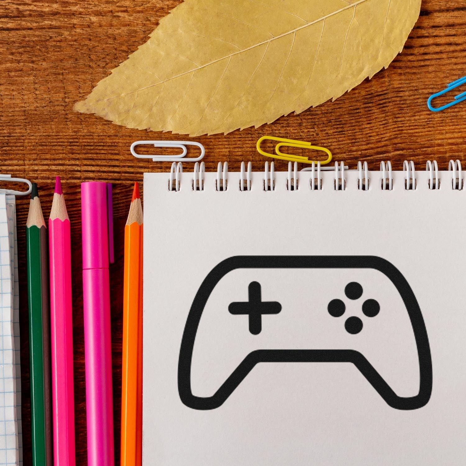 Self-Inking Game On! Reward Stamp on a notepad with a game controller icon, surrounded by colorful pencils and paperclips on a wooden desk. Perfect for teachers and parents to motivate kids.